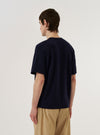 A model wears the Navy Merino Fine Knit Knit Tee against a plain, light-colored background. The navy blue knit t-shirt features a fine, smooth texture, a classic crew neckline, and subtle ribbed detailing at the sleeve cuffs and hem. The image focuses on the model from the chest up, highlighting the garment's drape and the quality of the merino wool fabric.