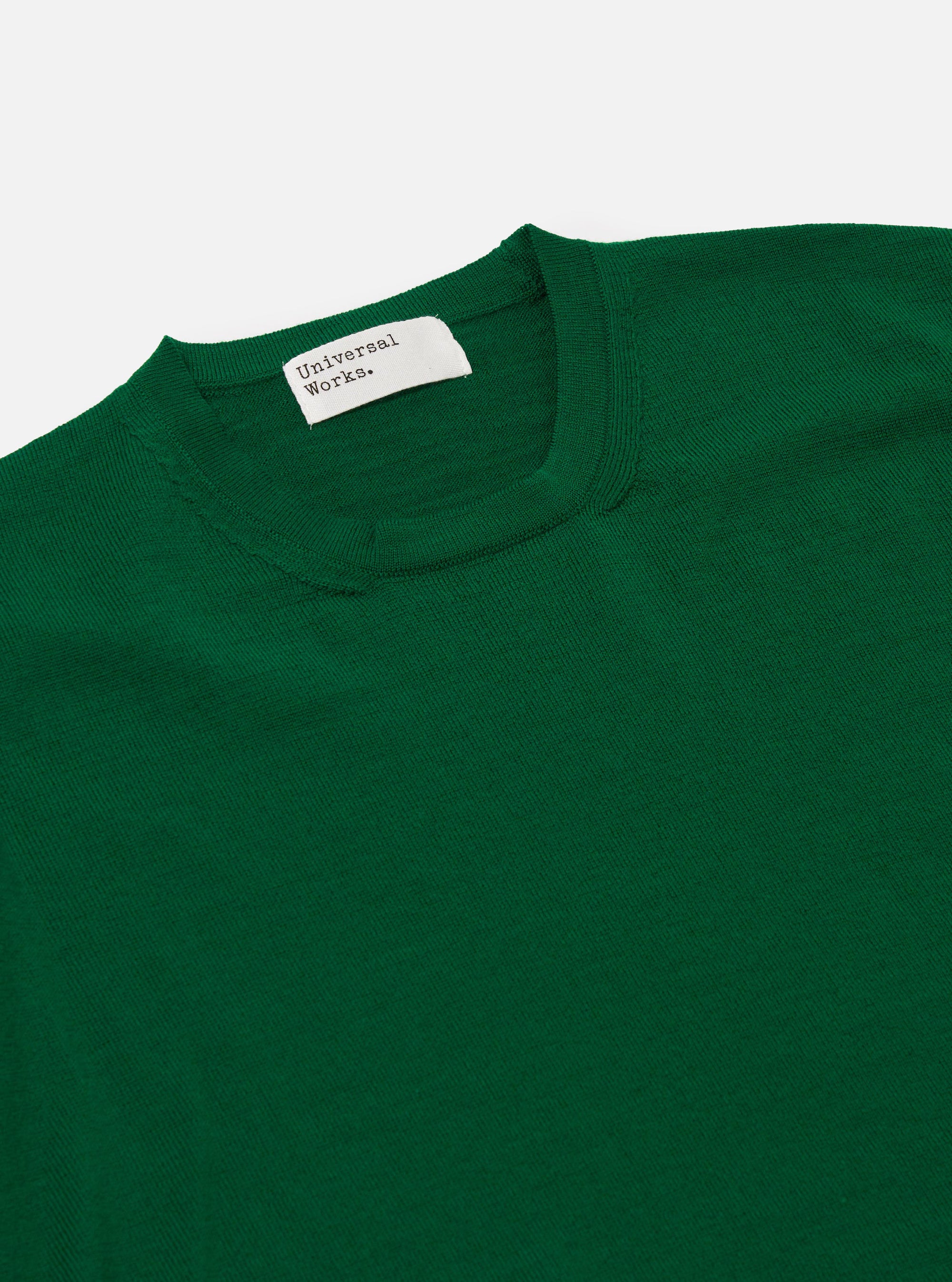 A model wears the Green Merino Fine Knit Knit Tee shown from a rear three-quarter view against a plain, light-colored background. The garment is a short-sleeved t-shirt made from a fine merino wool knit, featuring a ribbed crew neck, ribbed cuffs, and a ribbed hem. The fit is relaxed, and the model is shown from the waist up.