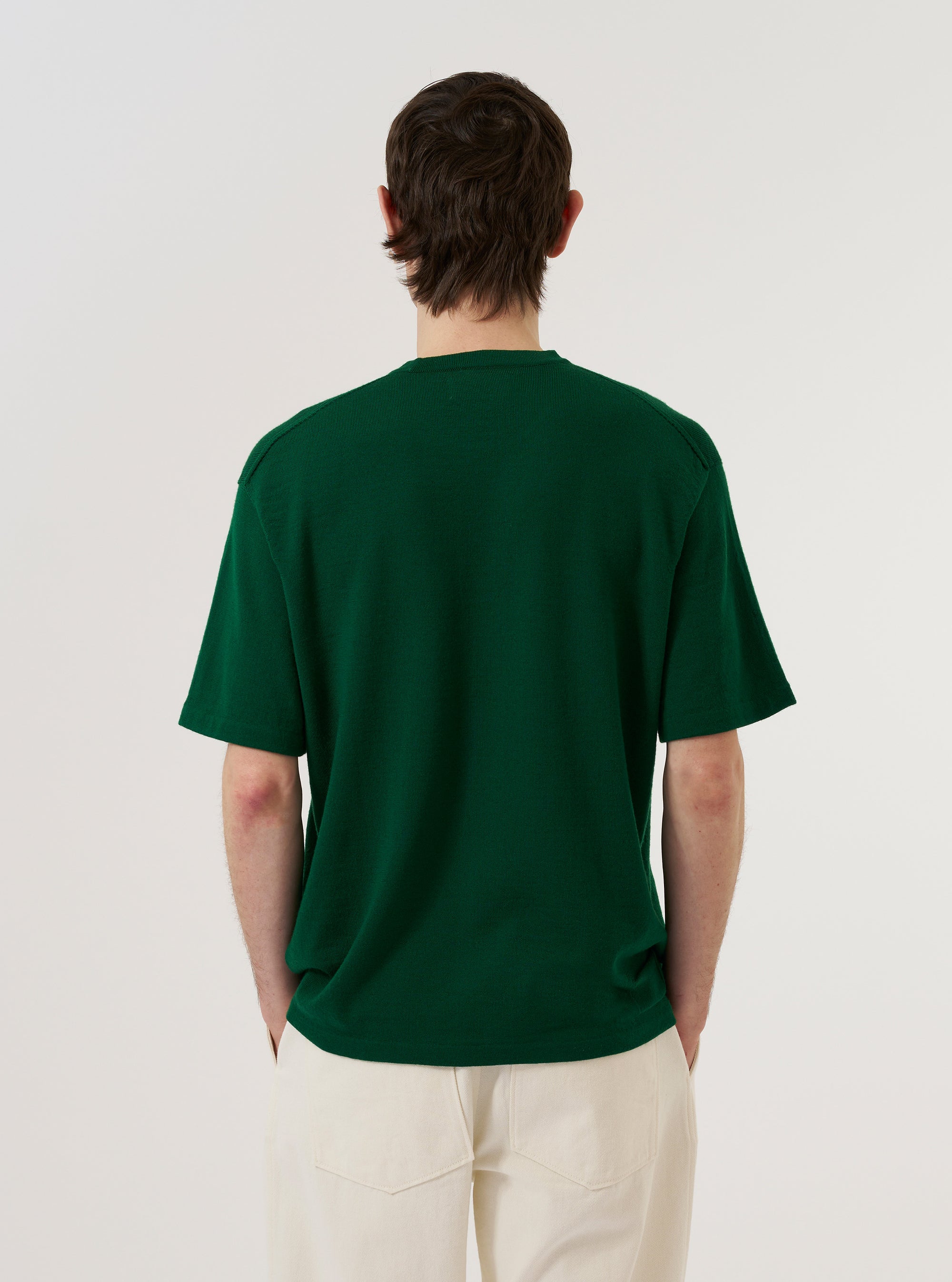 A model wears the Green Merino Fine Knit Knit Tee against a plain white background. The short-sleeved tee features a classic crew neck and a fine, lightweight texture with subtle ribbed detailing at the collar and cuffs. The model is shown from the waist up, standing with one hand in their pocket.