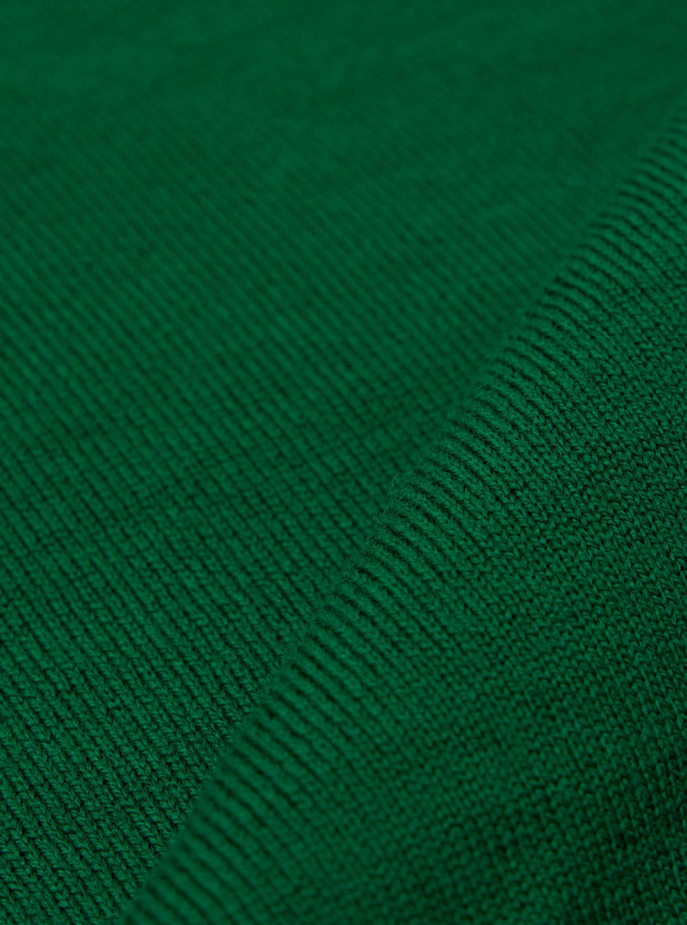 A close-up fabric swatch of the Green Merino Fine Knit Knit Tee highlights the material's fine gauge and soft, textured weave in a solid green color. The image focuses entirely on the fabric detail, and no model or mannequin is present.