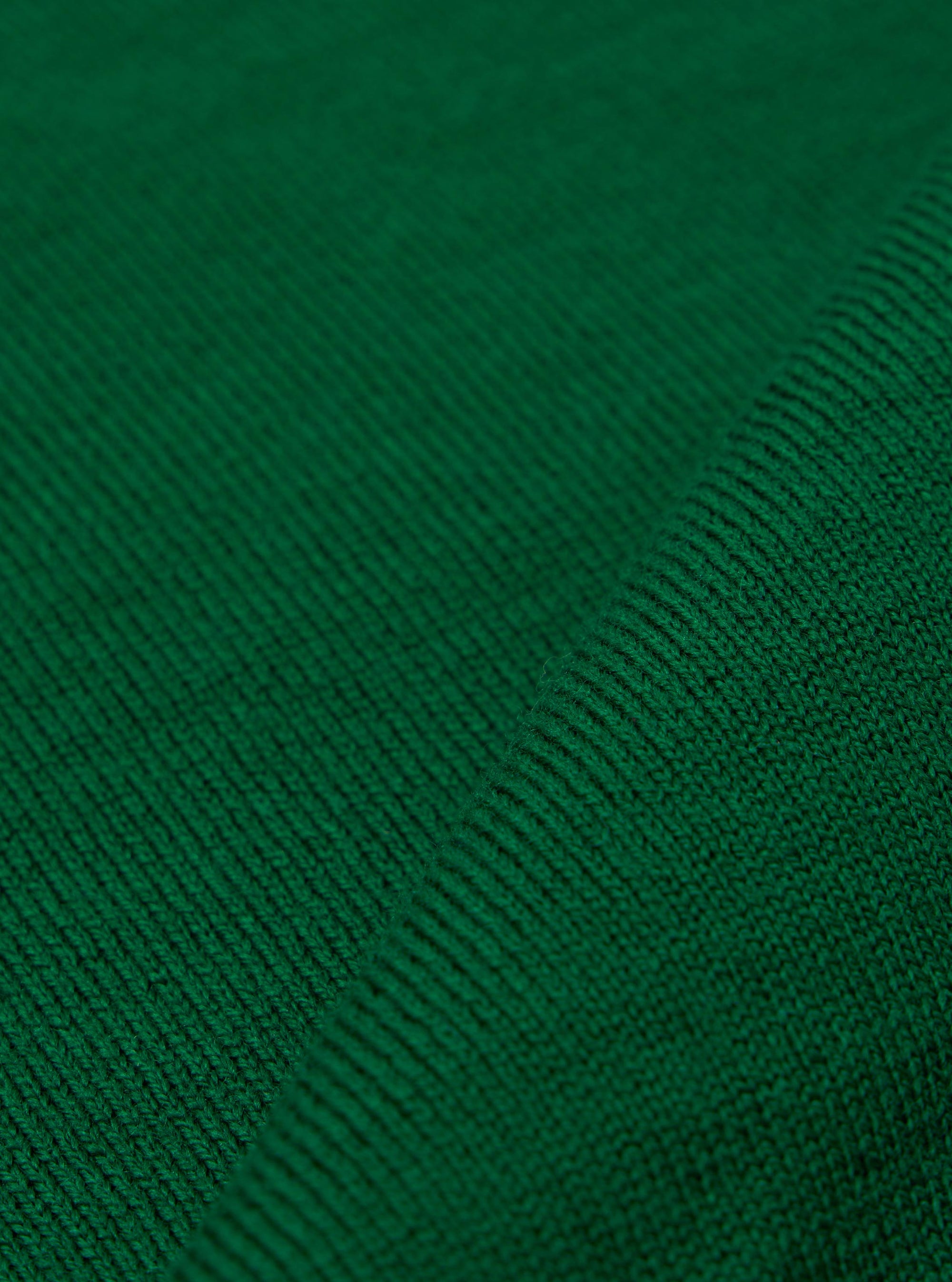 A close-up fabric swatch of the Green Merino Fine Knit Knit Tee highlights the material's fine gauge and soft, textured weave in a solid green color. The image focuses entirely on the fabric detail, and no model or mannequin is present.