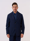 A model wears the Navy Derby Stripe Two Button Jacket over a light-colored shirt, standing against a plain, neutral background. The jacket features a two-button front closure, notched lapels, and two large patch pockets at the waist. The navy blue cotton fabric is detailed with a vertical stripe pattern.