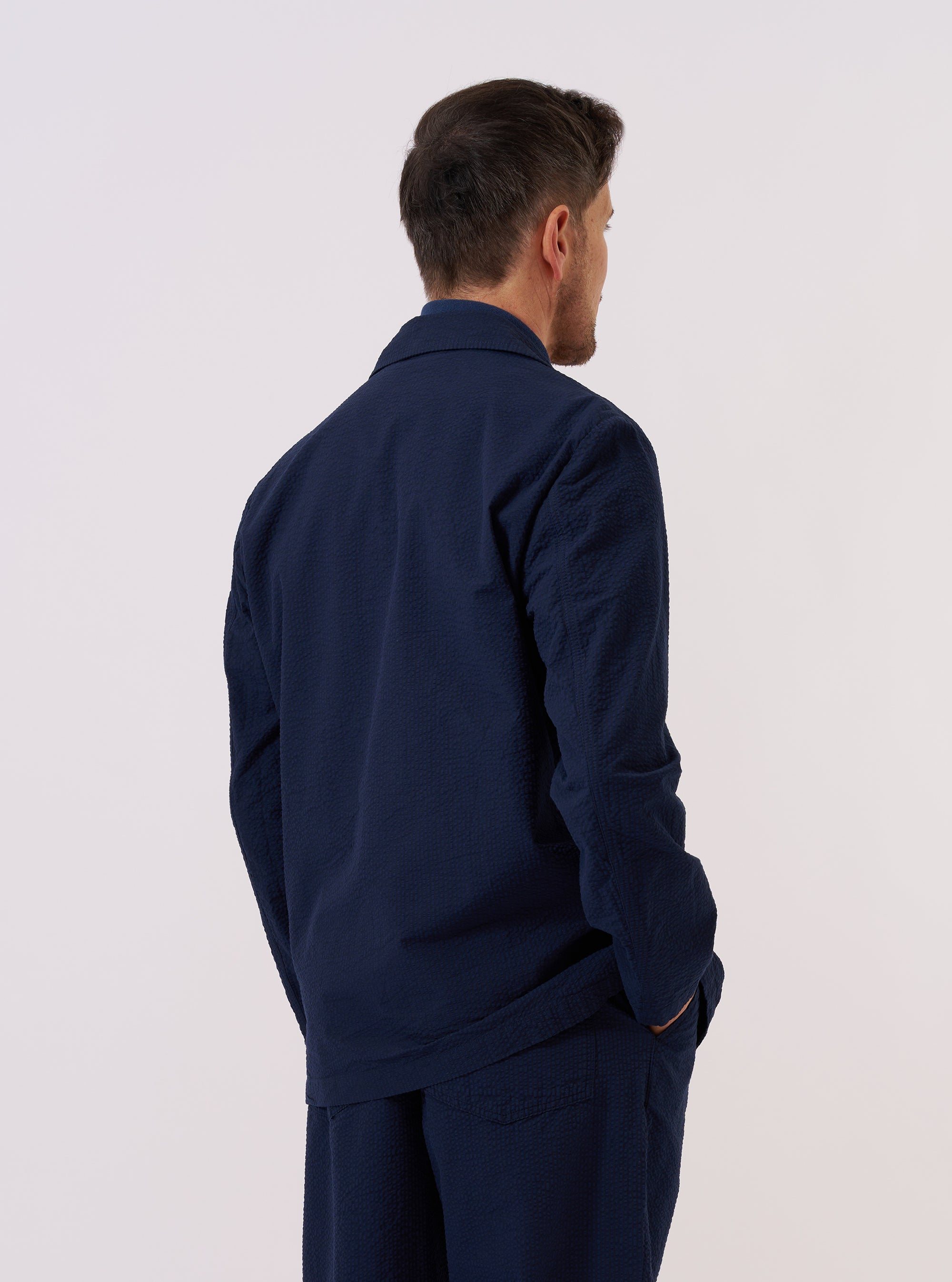 A model wears the Navy Derby Stripe Two Button Jacket over a light-colored top, shown from a side-angle view against a plain, neutral studio background. The jacket features a classic notched lapel, two-button front closure, and a visible vertical stripe pattern on the navy cotton fabric. The image highlights the tailored fit and the structured shoulder of the garment.