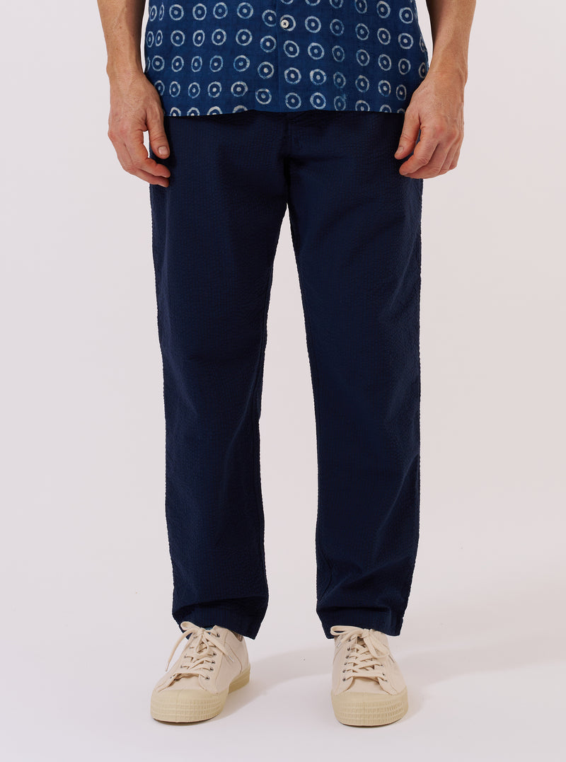 A model wears the Navy Derby Stripe Military Chino against a plain white background. The image shows a front view of the trousers, highlighting the slim, straight-leg fit and the navy blue fabric with thin vertical white derby stripes. The model is wearing the chinos with a tucked-in shirt to show the waistband and button closure.