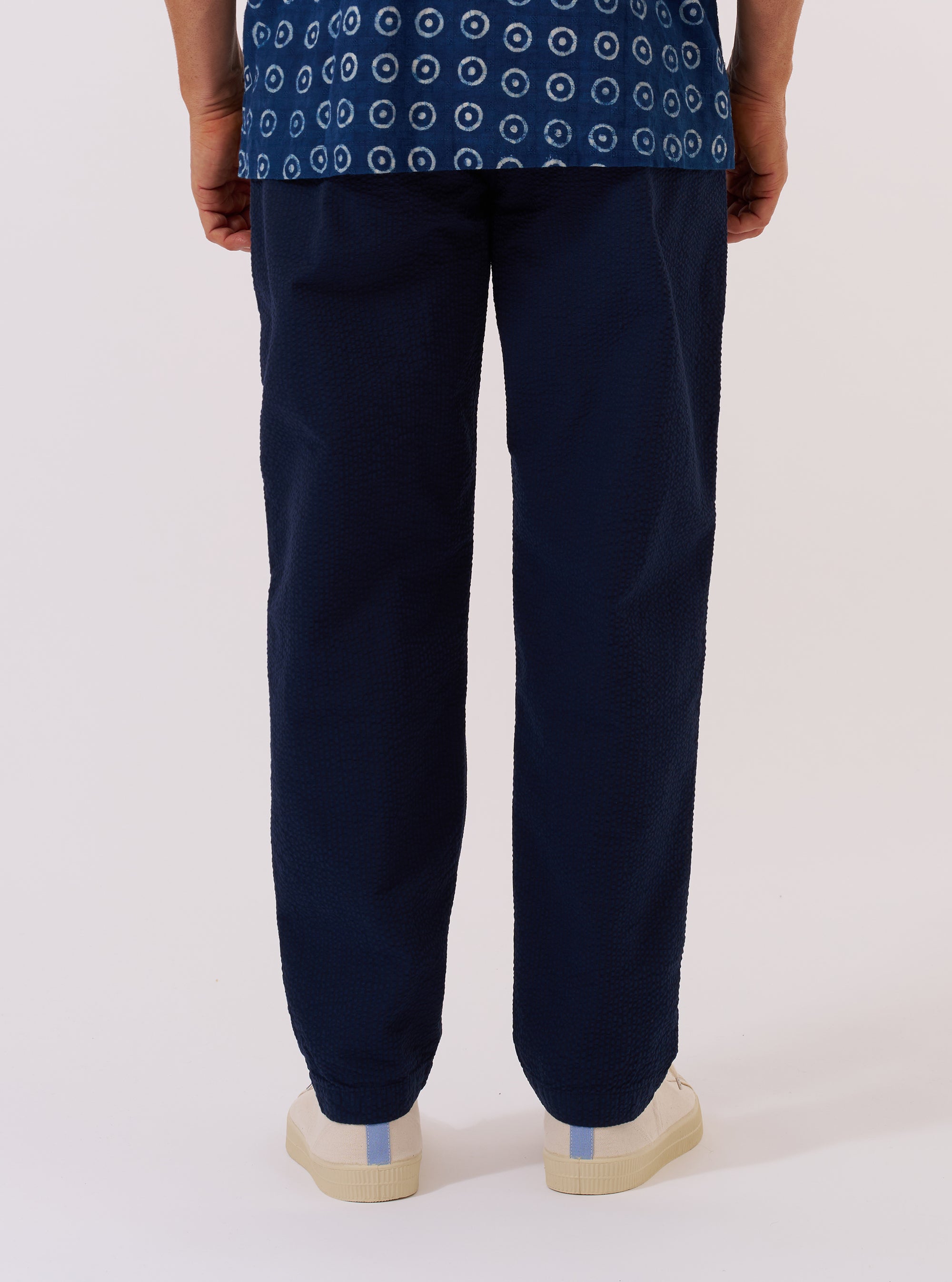 A model wears the Navy Derby Stripe Military Chino, shown from a rear angle against a plain white background. The trousers feature a straight-leg fit, two button-down rear patch pockets, and a distinct vertical stripe pattern. The cotton fabric is a deep navy with contrasting light stripes. The model is standing, highlighting the garment's silhouette and pocket placement.