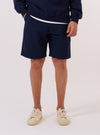 A model wears the Navy Derby Stripe Clean Short against a plain, neutral background. The shorts feature a bold vertical navy and off-white stripe pattern, an elasticated waistband, and a relaxed, straight-leg fit. The image highlights the garment's pattern and side pocket placement as the model stands facing forward.