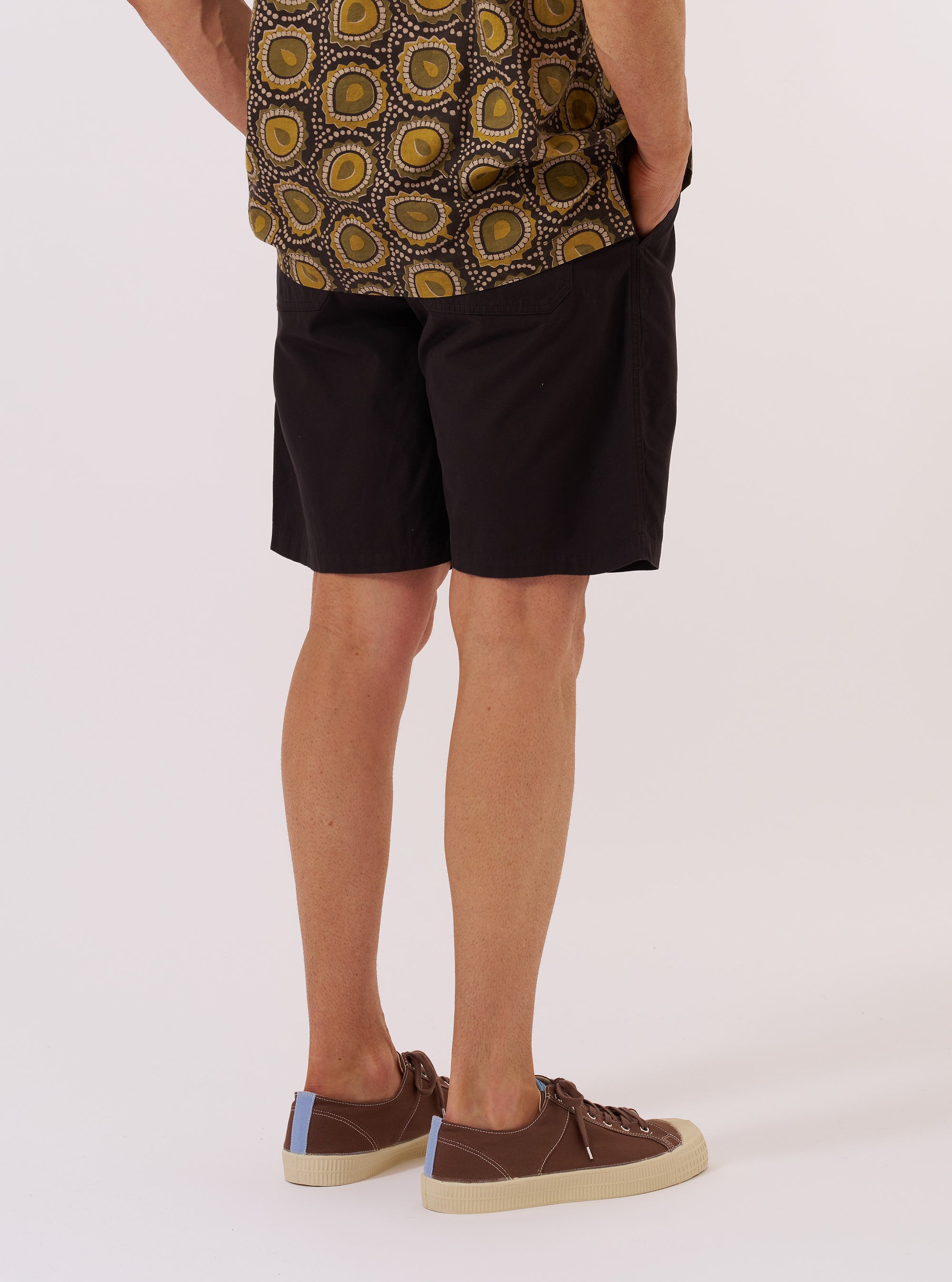 A model is wearing the Licorice Summer Canvas Clean Short, shown from the waist down against a plain, neutral background. The shorts feature a relaxed fit and are made from a dark brown licorice-colored cotton canvas. The garment has a clean-cut appearance with visible side pockets and a structured waistband.