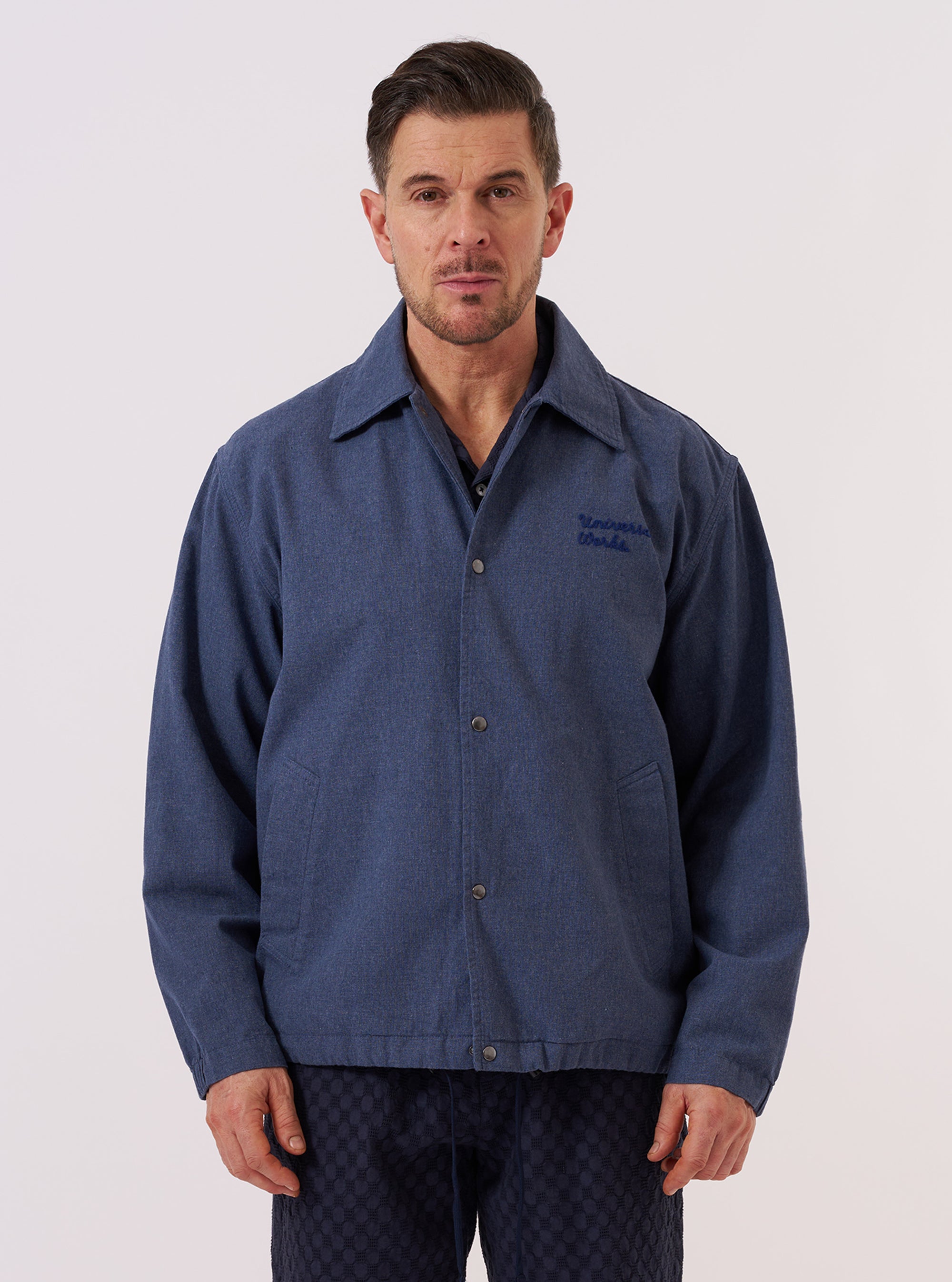 A model wears the Blue Sintra Recycled Cotton Head Coach Jacket over a light-colored shirt, standing against a plain, neutral background. The jacket features a pointed collar, a central snap-button closure, and two large slanted welt pockets at the waist. The blue fabric has a smooth, matte finish.
