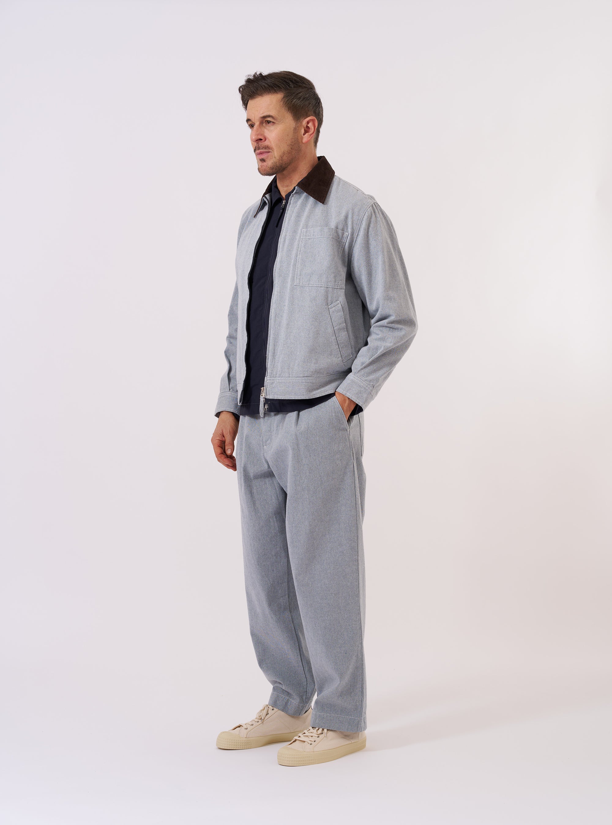 The Light Indigo Jean Cloth Reworked Rose Bowl Jacket is shown as a flat lay against a white background. The jacket features a boxy fit with a full-length silver double-ended zip front, a structured collar, and buttoned adjustable cuffs. One large patch pocket is visible on the chest, along with two side welt pockets. The light indigo fabric is made from a reworked jean cloth with visible panelled construction and tonal stitching.