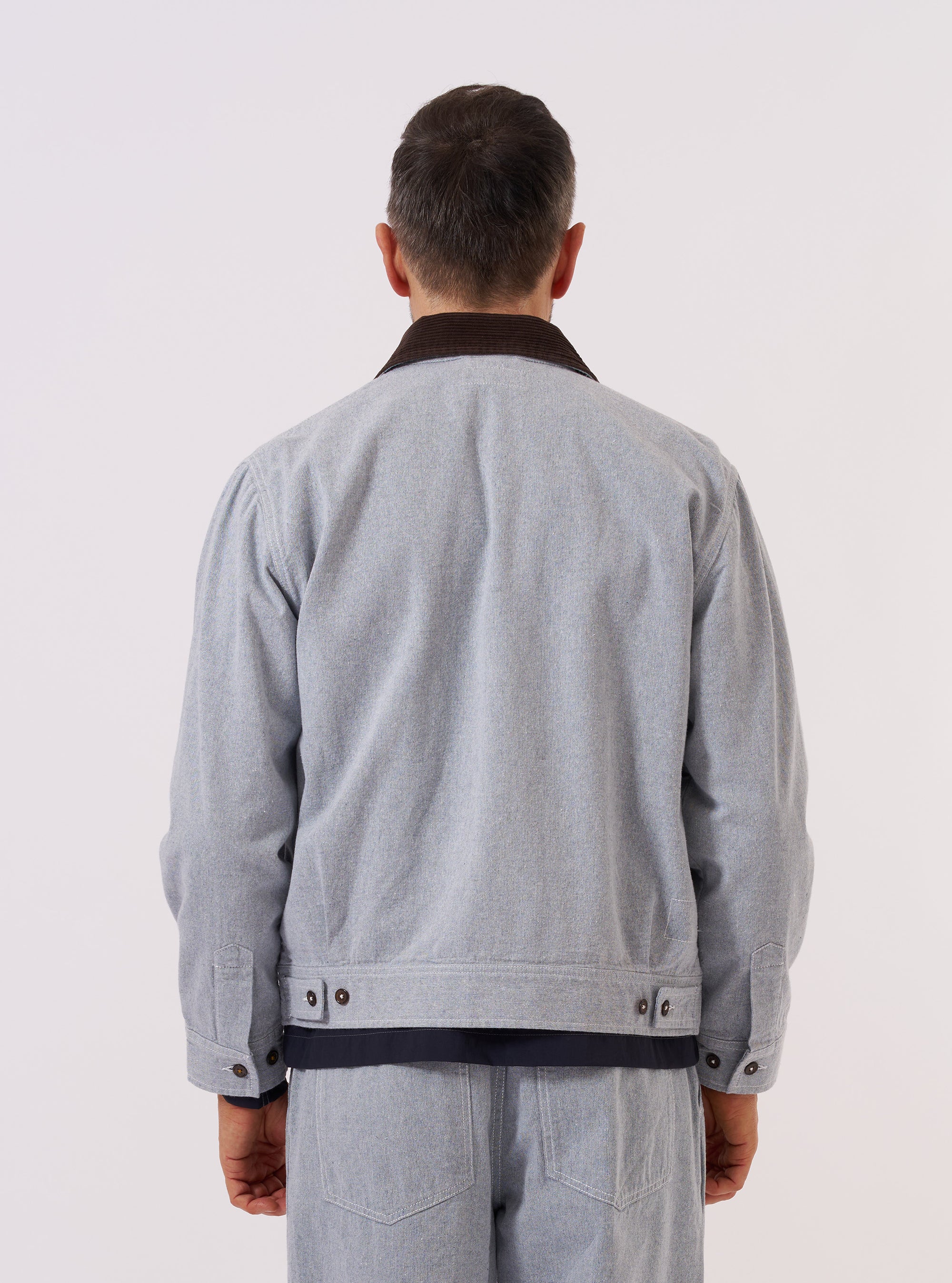 A model wears the Light Indigo Jean Cloth Reworked Rose Bowl Jacket, shown from a rear three-quarter angle against a plain white background. The jacket features a relaxed fit with a straight hem, a structured collar, and long sleeves. The light indigo organic cotton fabric has a visible textured grain, and the back is constructed with a clean, one-piece panel. No mannequin is present in the image.