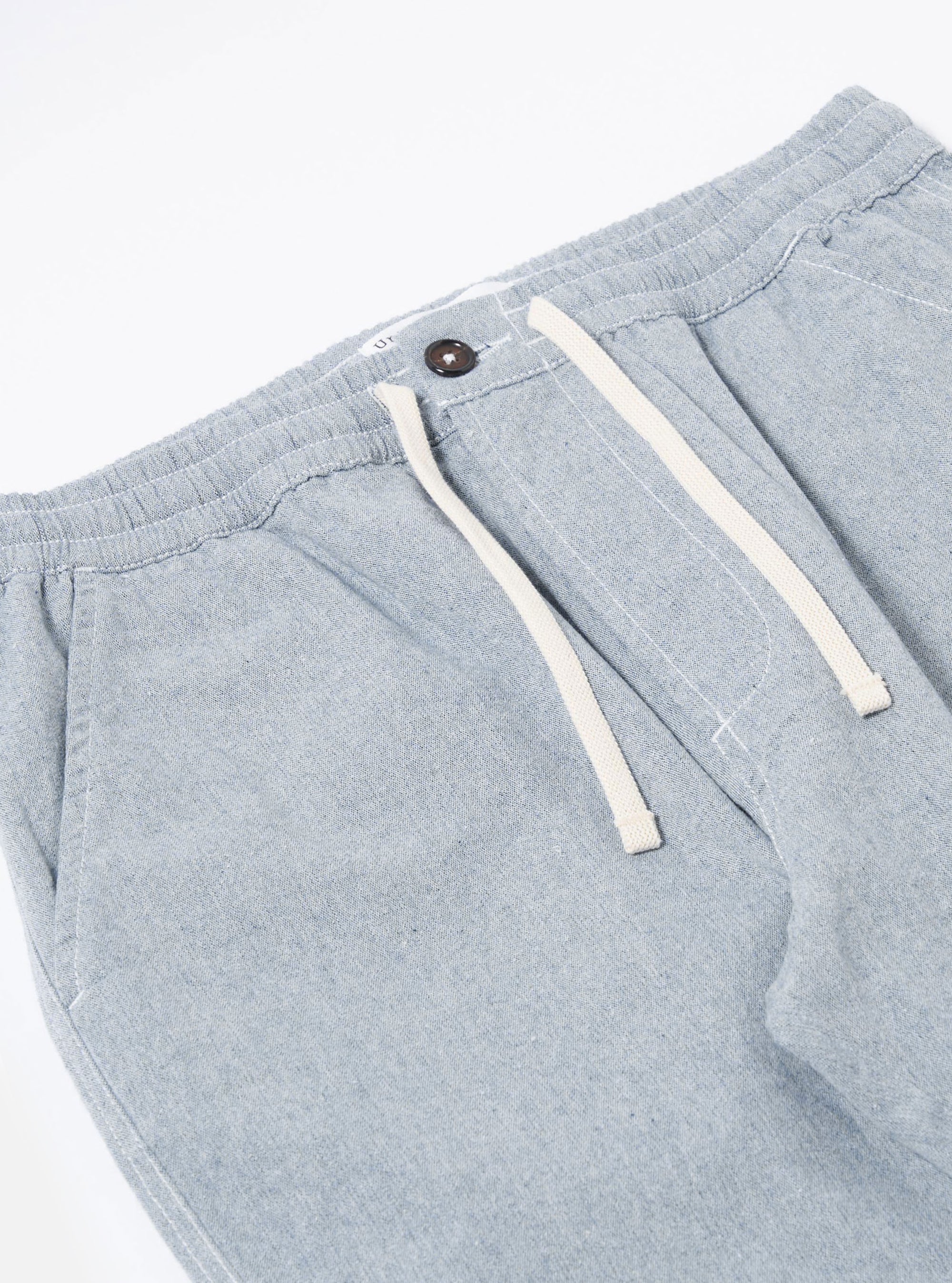 The Light Indigo Jean Cloth Reworked Hi Water Trouser is shown from a rear angle as a flat lay against a plain white background. The image highlights the two back patch pockets and the elasticated drawstring waistband. The light indigo organic cotton fabric features a visible textured weave and a slightly cropped leg length.
