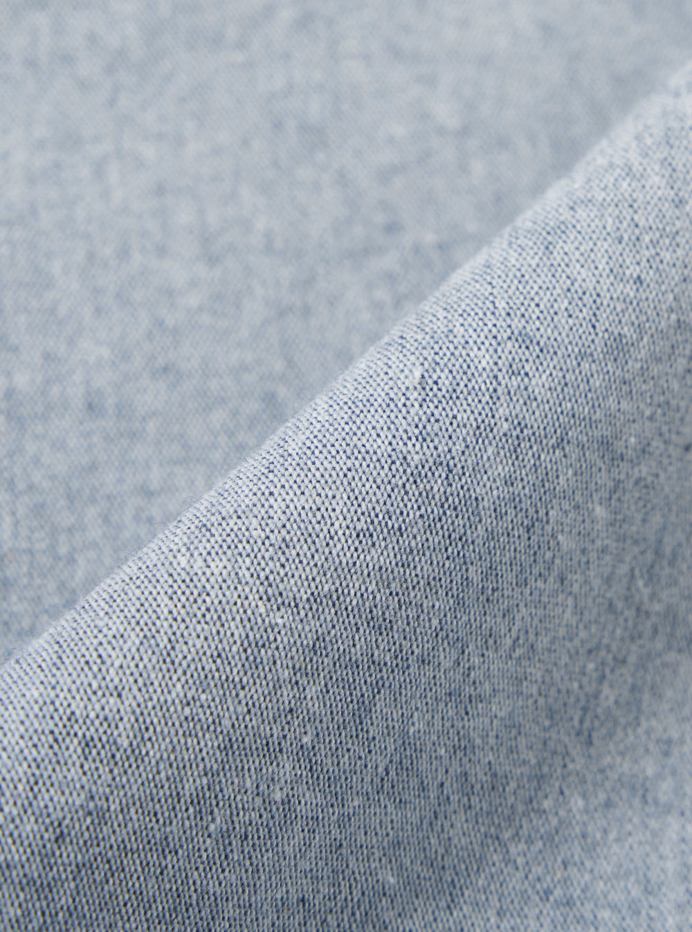 A close-up fabric swatch of the Light Indigo Jean Cloth Reworked Hi Water Trouser displays a light blue indigo denim with a subtle textured weave. The organic cotton fabric is shown in detail, filling the frame to highlight the material's color and construction.