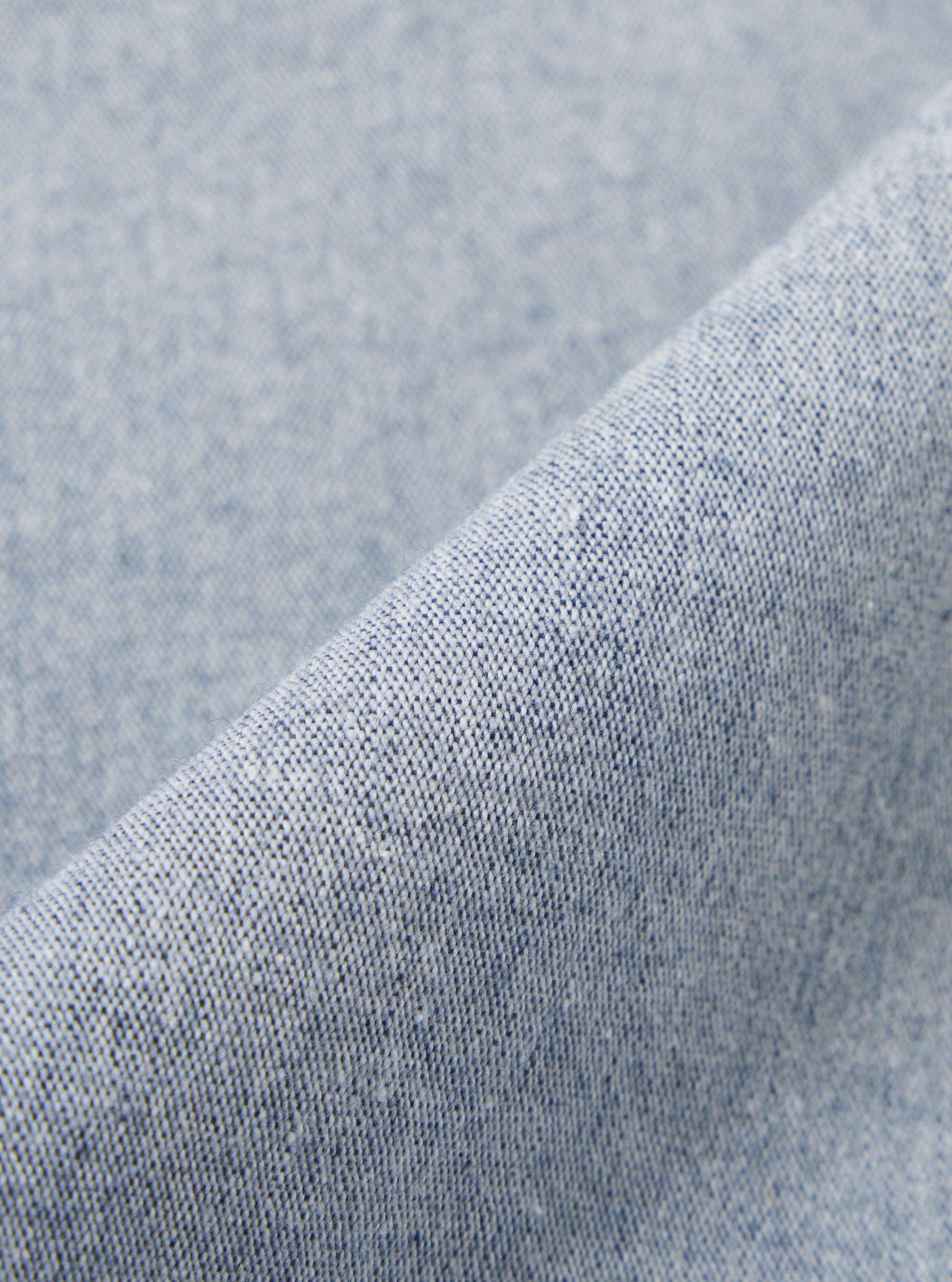 A close-up fabric swatch of the Light Indigo Jean Cloth Reworked Hi Water Trouser displays a light blue indigo denim with a subtle textured weave. The organic cotton fabric is shown in detail, filling the frame to highlight the material's color and construction.