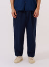 A model wears the Navy Derby Stripe Oxford Pant against a plain white background. The trousers feature a loose, wide-leg fit with a pleated front and a drawstring waistband. The navy blue cotton fabric has a prominent vertical white stripe pattern throughout. The model is shown from the waist down, paired with a light-colored shirt and dark shoes.