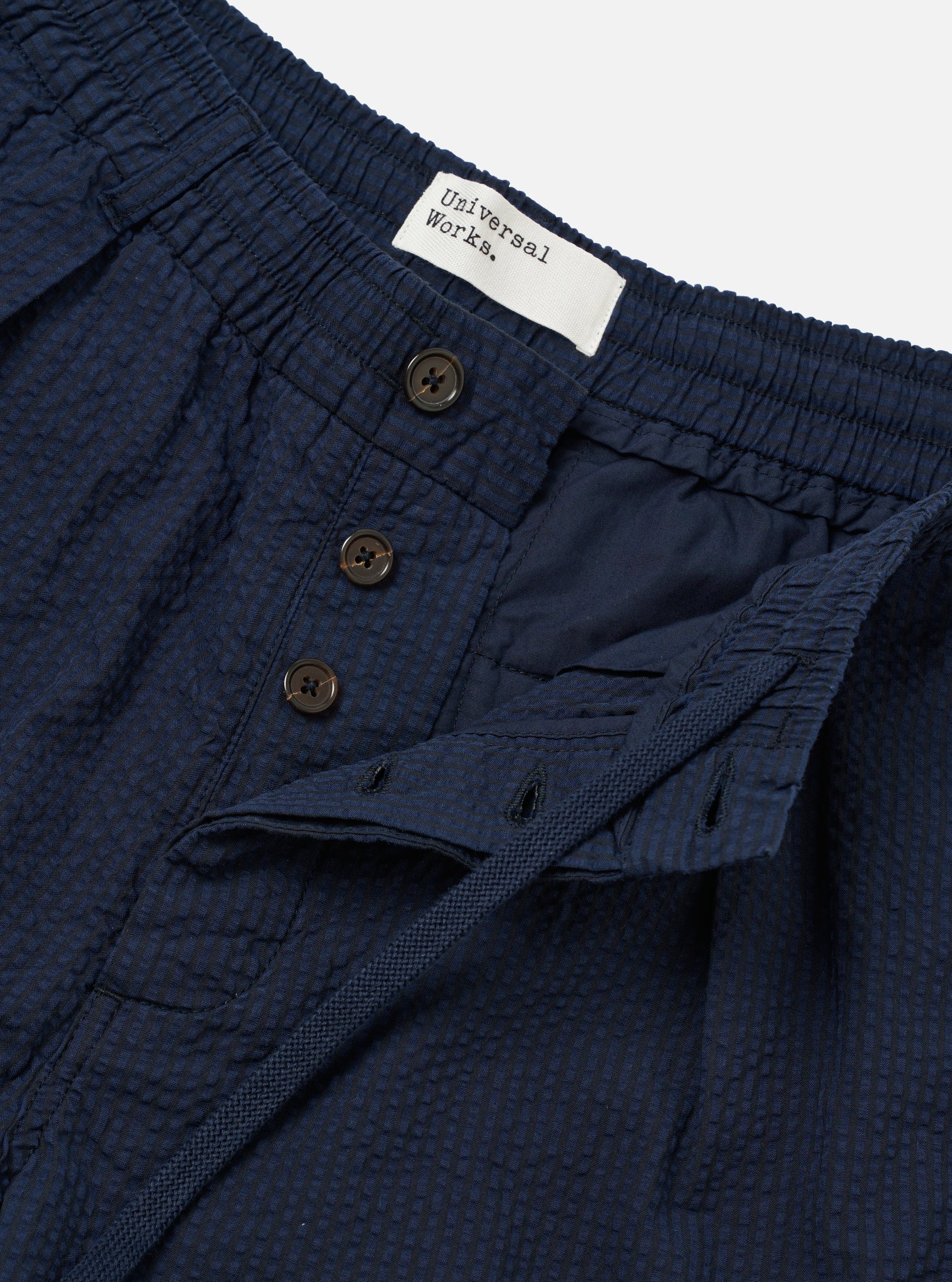 The Navy Derby Stripe Oxford Pant is shown in a detail view as a flat lay against a plain white background. The image highlights the construction of the waistband, featuring an internal drawstring and front pleats. The cotton fabric displays a distinct navy and white vertical derby stripe pattern with visible textured stitching.