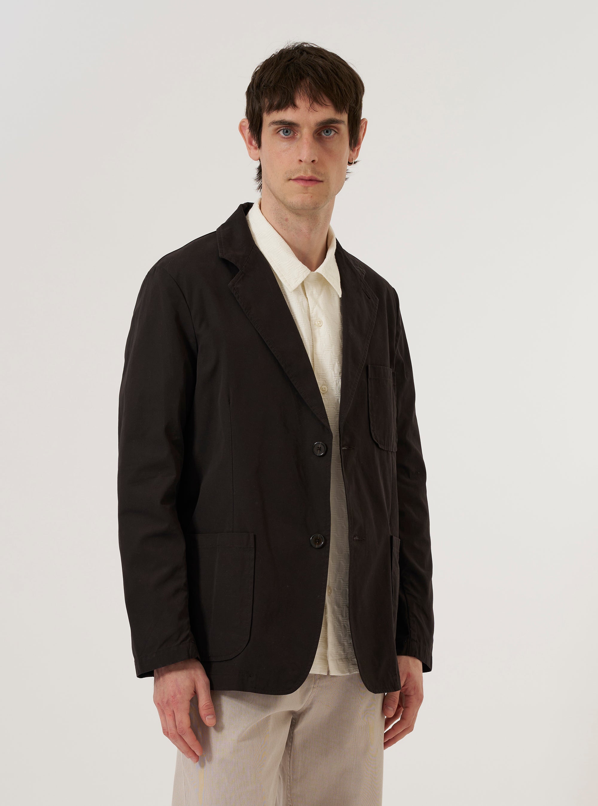A model wears the Licorice Summer Canvas Two Button Jacket over a dark top, standing against a plain white background. The brown jacket features a loose fit with a two-button front closure, wide notched lapels, and large patch pockets at the waist. The summer canvas fabric has a visible matte texture and a structured appearance.