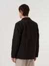 A model wears the Licorice Summer Canvas Two Button Jacket over a light-colored shirt, shown from a rear three-quarter angle against a plain white studio background. The jacket features a structured back, a single rear vent, and a relaxed tailored fit. The licorice brown fabric has a visible canvas texture.