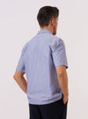 A model is wearing the Blue Oxford Shirting Road Trip Shirt over a light-colored t-shirt against a plain white background. The image, shown from the waist up, captures the model from a three-quarter rear angle, highlighting the shirt's relaxed fit, short sleeves, and the smooth texture of the blue Oxford fabric.