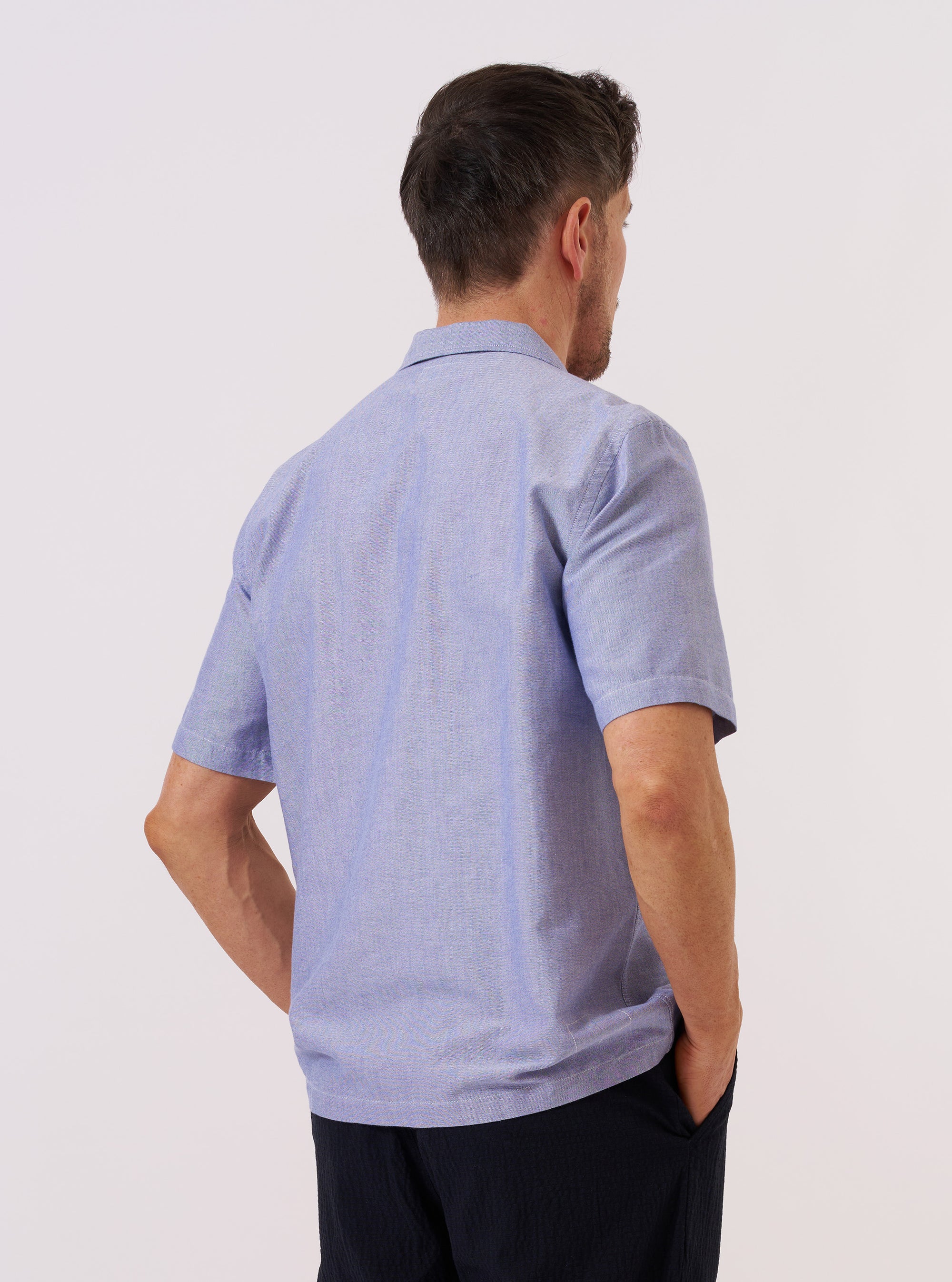 A model is wearing the Blue Oxford Shirting Road Trip Shirt over a light-colored t-shirt against a plain white background. The image, shown from the waist up, captures the model from a three-quarter rear angle, highlighting the shirt's relaxed fit, short sleeves, and the smooth texture of the blue Oxford fabric.