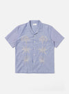 The Blue Oxford Shirting Road Trip Shirt is shown as a flat lay against a white background. The shirt features a camp collar, a central button-down front with tonal buttons, and a straight hem. The light blue cotton Oxford fabric has a visible weave, and a single patch pocket is located on the left chest.