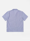 The Blue Oxford Shirting Road Trip Shirt is shown from the back as it is worn by a model against a neutral studio background. The shirt features a clean, horizontal yoke across the shoulders and a straight-cut hem. The model has their hands in their pockets, highlighting the shirt's relaxed fit and the textured appearance of the blue cotton oxford fabric. No mannequin is present in the image.