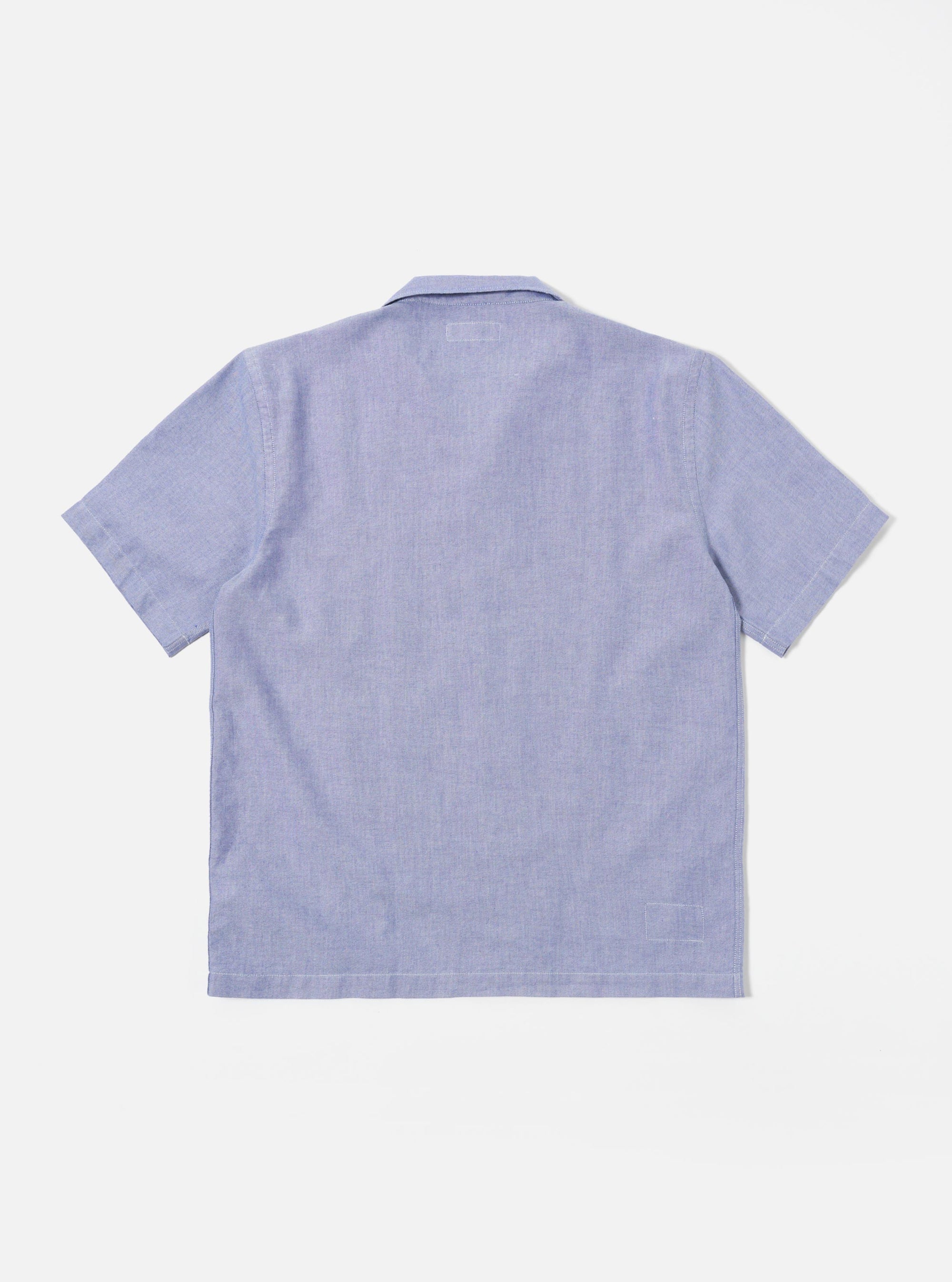 The Blue Oxford Shirting Road Trip Shirt is shown from the back as it is worn by a model against a neutral studio background. The shirt features a clean, horizontal yoke across the shoulders and a straight-cut hem. The model has their hands in their pockets, highlighting the shirt's relaxed fit and the textured appearance of the blue cotton oxford fabric. No mannequin is present in the image.