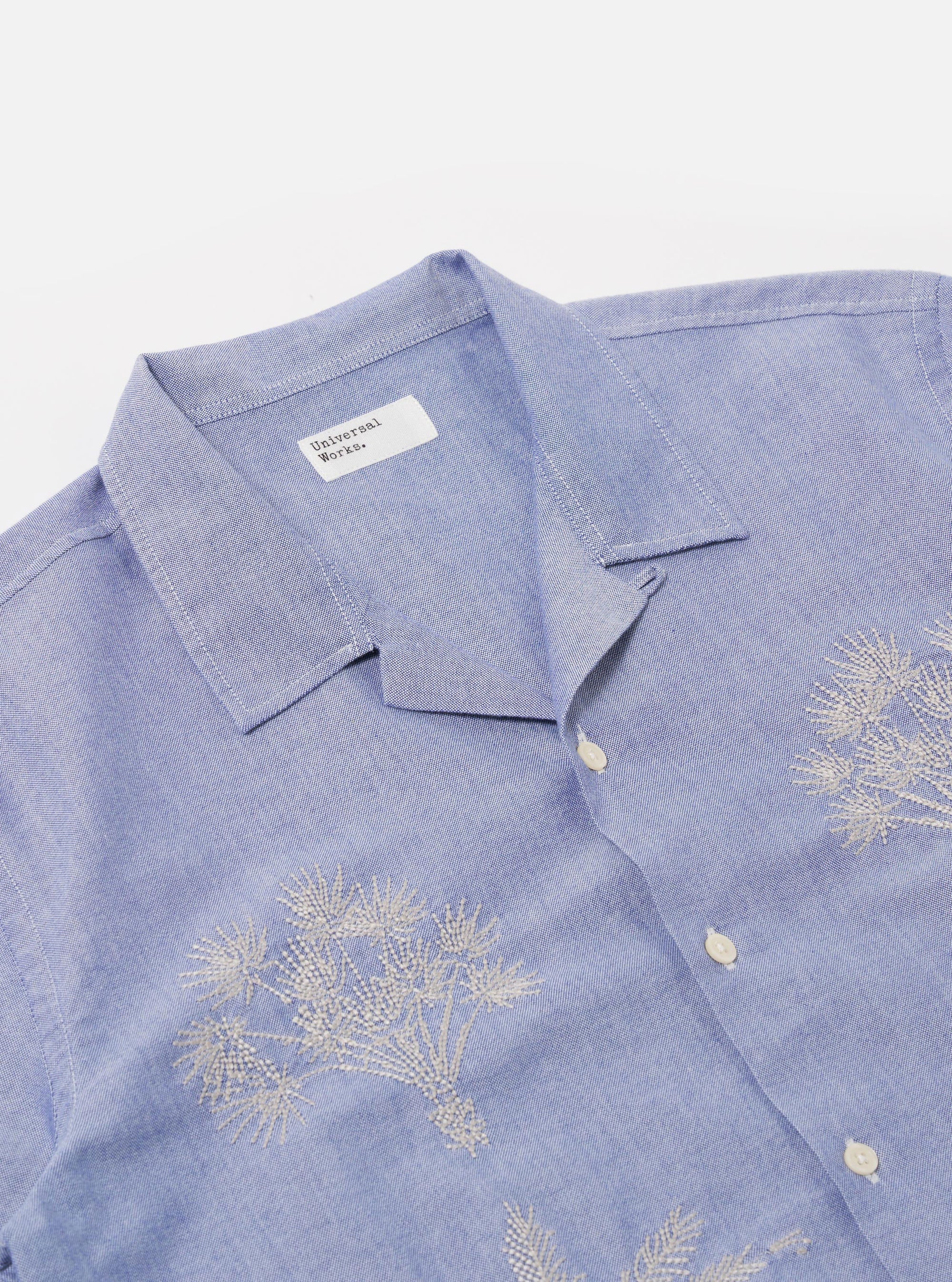 The Blue Oxford Shirting Road Trip Shirt is shown as a flat lay against a white background. The shirt is a light blue, short-sleeved button-up with a regular fit and a straight hem. It features a single-piece loop-close collar, a single chest patch pocket, and tonal buttons.