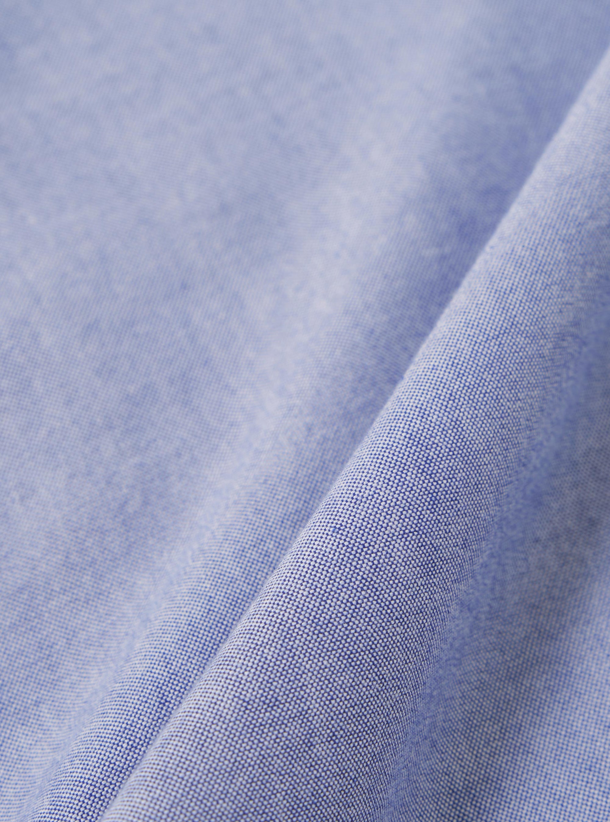 A close-up fabric swatch of the Blue Oxford Shirting Road Trip Shirt shows the detailed texture of the blue Oxford cotton material. The image displays the characteristic weave of blue and white threads.