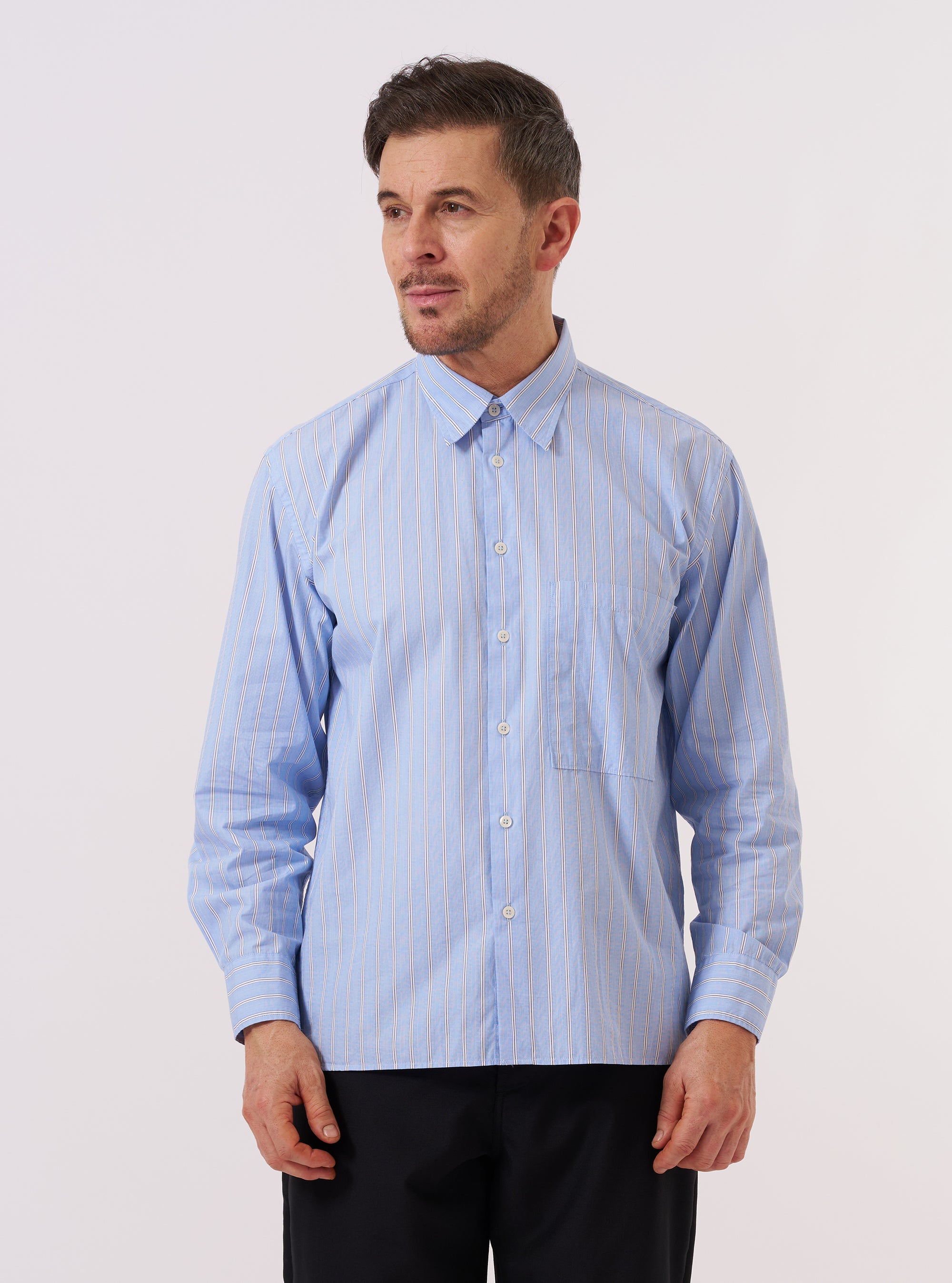 A model wears the Blue Mc Stripe Square Pocket Shirt against a plain, light grey background. The shirt features a blue and white vertical stripe pattern, a classic button-down front, a structured collar, and two large square patch pockets at the chest. The model is shown from a front-facing angle, wearing the shirt buttoned up with the sleeves down.