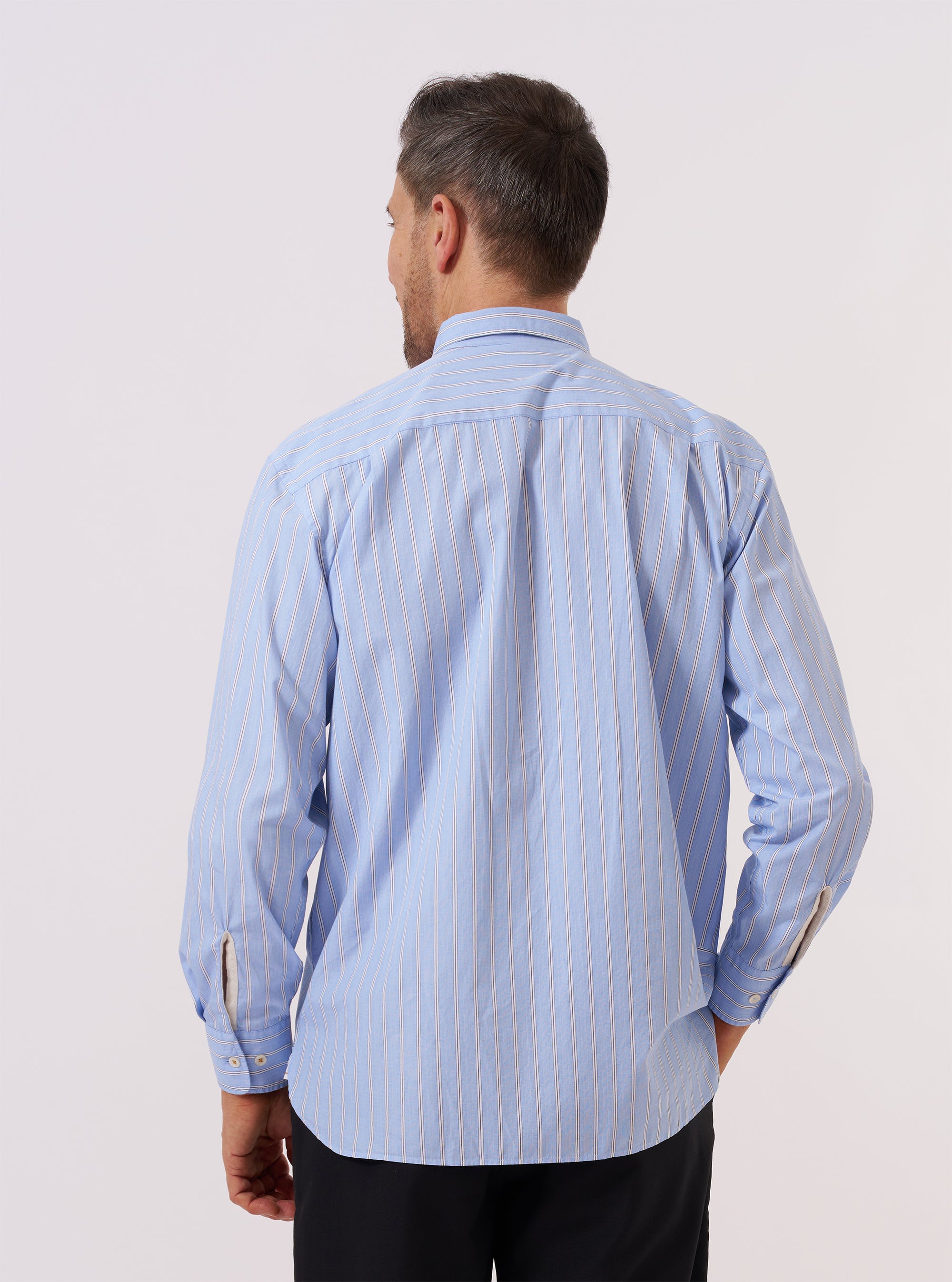 A model is wearing the Blue Mc Stripe Square Pocket Shirt, shown from the back against a plain white background. The shirt features a classic blue and white vertical stripe pattern, a straight hem, and a relaxed fit. The model is wearing the shirt over a light-colored top and dark trousers.