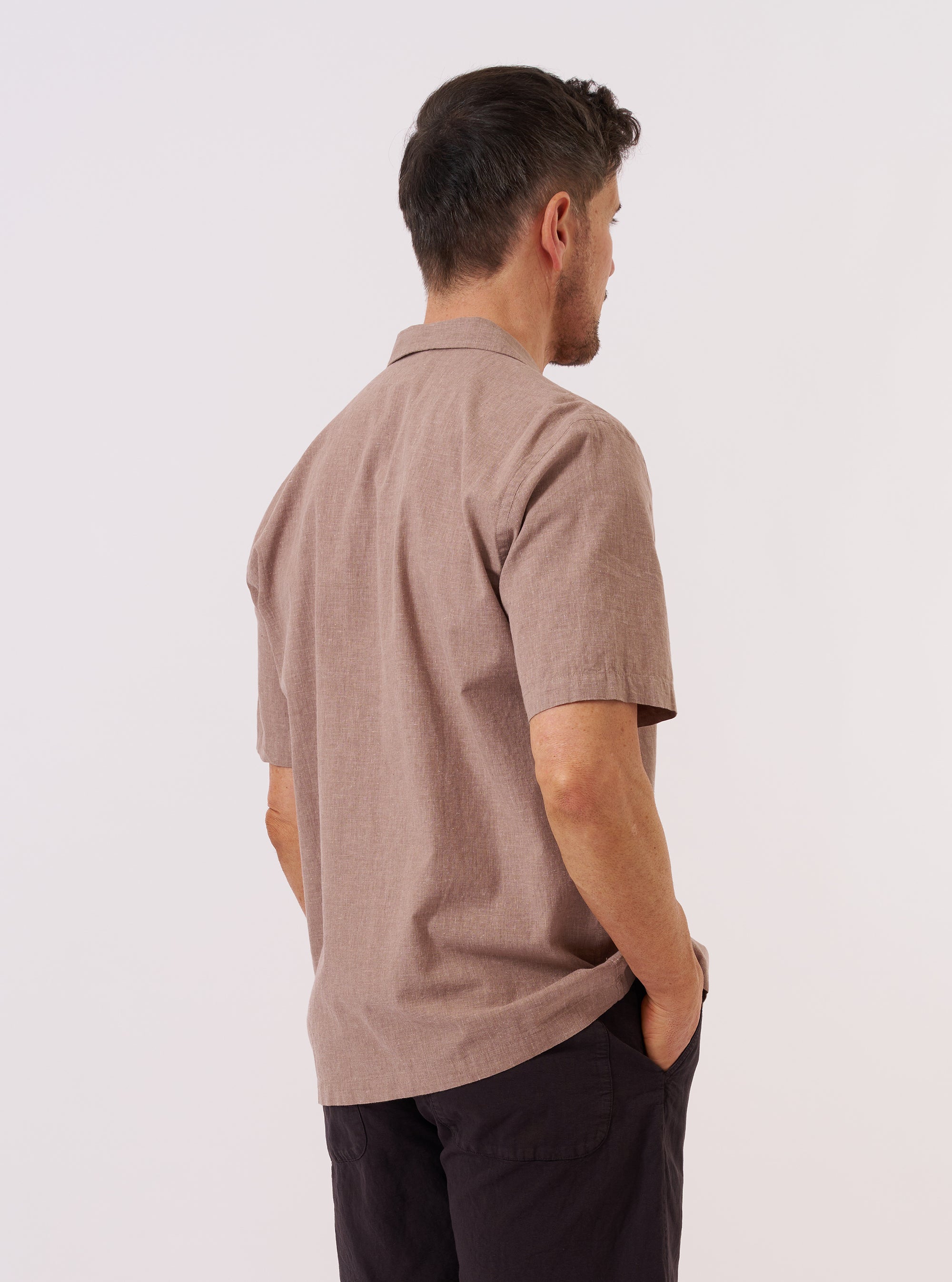 A model is wearing the Brown Sido Linen Mix Road Shirt over a white t-shirt, shown from the back and side as they walk against a plain white background. The image highlights the shirt's relaxed fit, straight hem, and short sleeves. The textured brown linen-blend fabric is clearly visible.