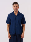 The Indigo Lincot Denim Road Shirt is worn by a model standing against a plain white background. The short-sleeve shirt is shown from the front, featuring a single chest patch pocket and a one-piece collar worn open. The model pairs the indigo-colored shirt with light-colored trousers.