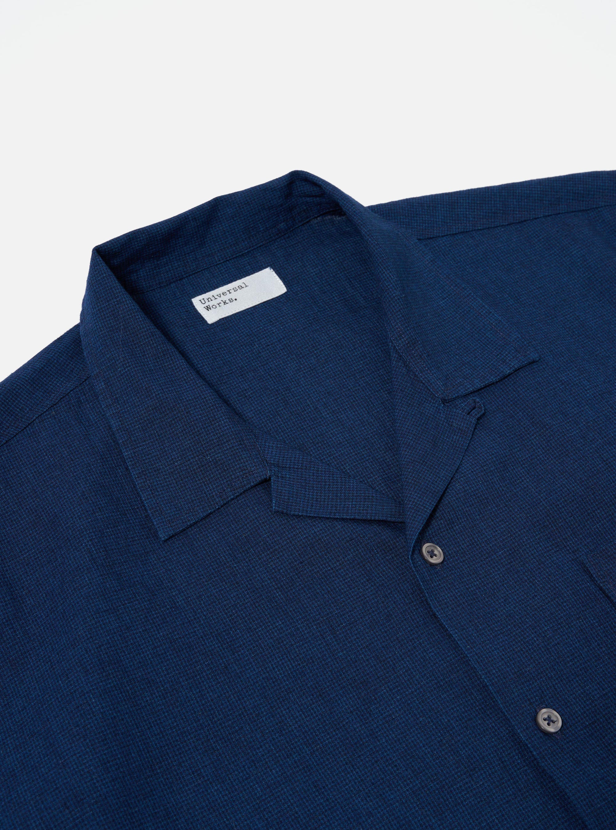 The Indigo Lincot Denim Road Shirt is worn by a model from a rear three-quarter angle against a plain, light-colored background. The shirt features a straight hem, short sleeves, and a structured one-piece collar. The indigo-dyed denim fabric has a visible textured weave. The model is also wearing light-colored trousers, and the shirt is worn untucked.