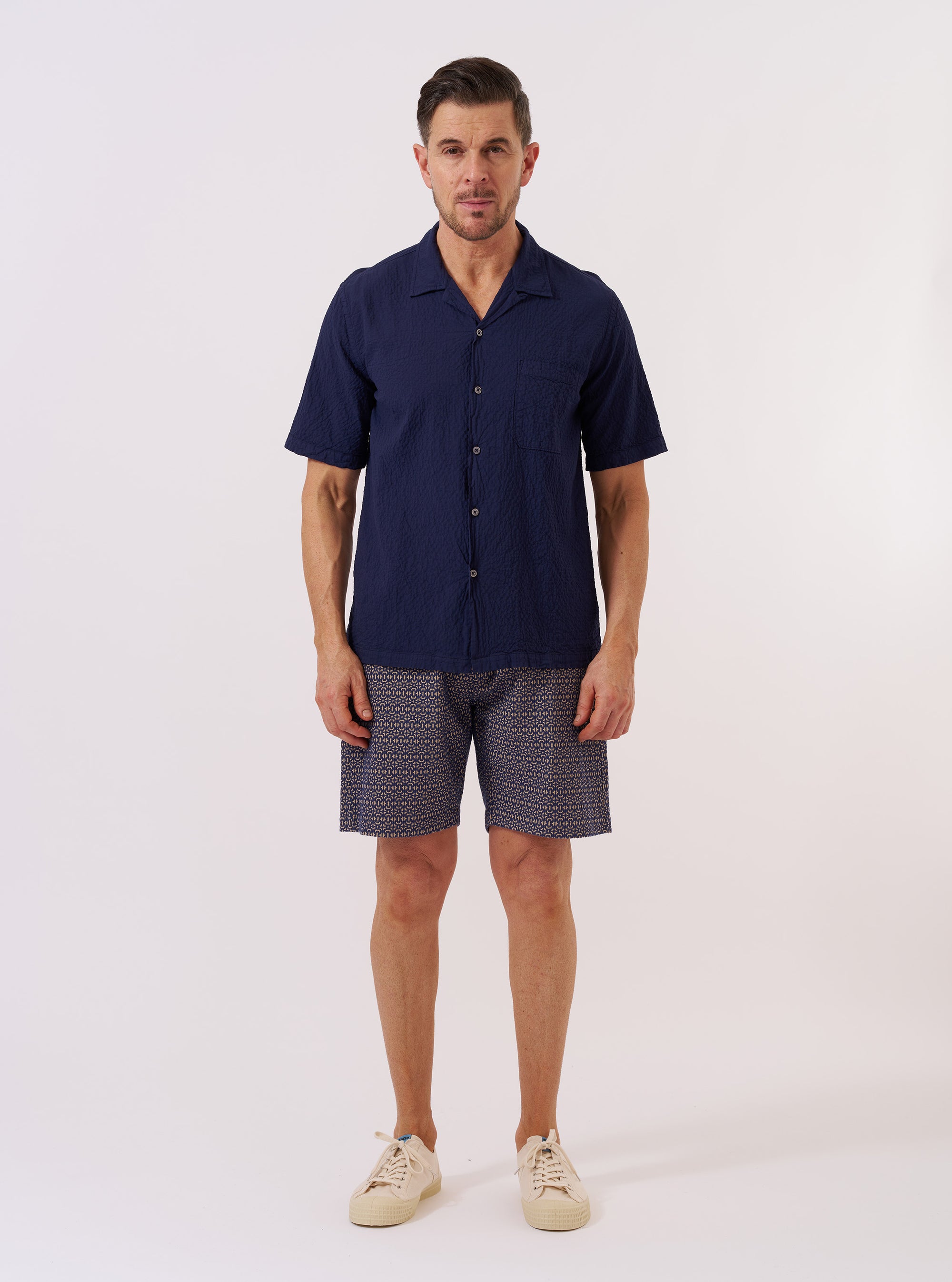 The Navy Arten Seersucker Road Shirt is displayed as a flat lay against a plain white background. The shirt features a short-sleeved design with a camp collar, a full button-down front, and a single patch pocket on the left chest. The navy organic cotton fabric has a distinct puckered seersucker texture throughout.