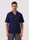 A model wears the Navy Arten Seersucker Road Shirt against a plain, light grey background. The short-sleeved shirt features a one-piece camp collar worn open, a central button-down front, and a single patch pocket on the left chest. The dark navy organic cotton fabric has a distinct puckered seersucker texture throughout. The model is shown from the waist up, highlighting the shirt's regular fit and straight hem.