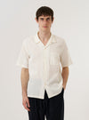 A model wears the Ecru Arten Seersucker Road Shirt against a plain, neutral background. The shirt features a camp collar, a central button-down front, and a single chest pocket on the left side. The ecru-colored organic cotton fabric has a distinct puckered seersucker texture. The model is shown from the front with the shirt tucked into dark trousers.