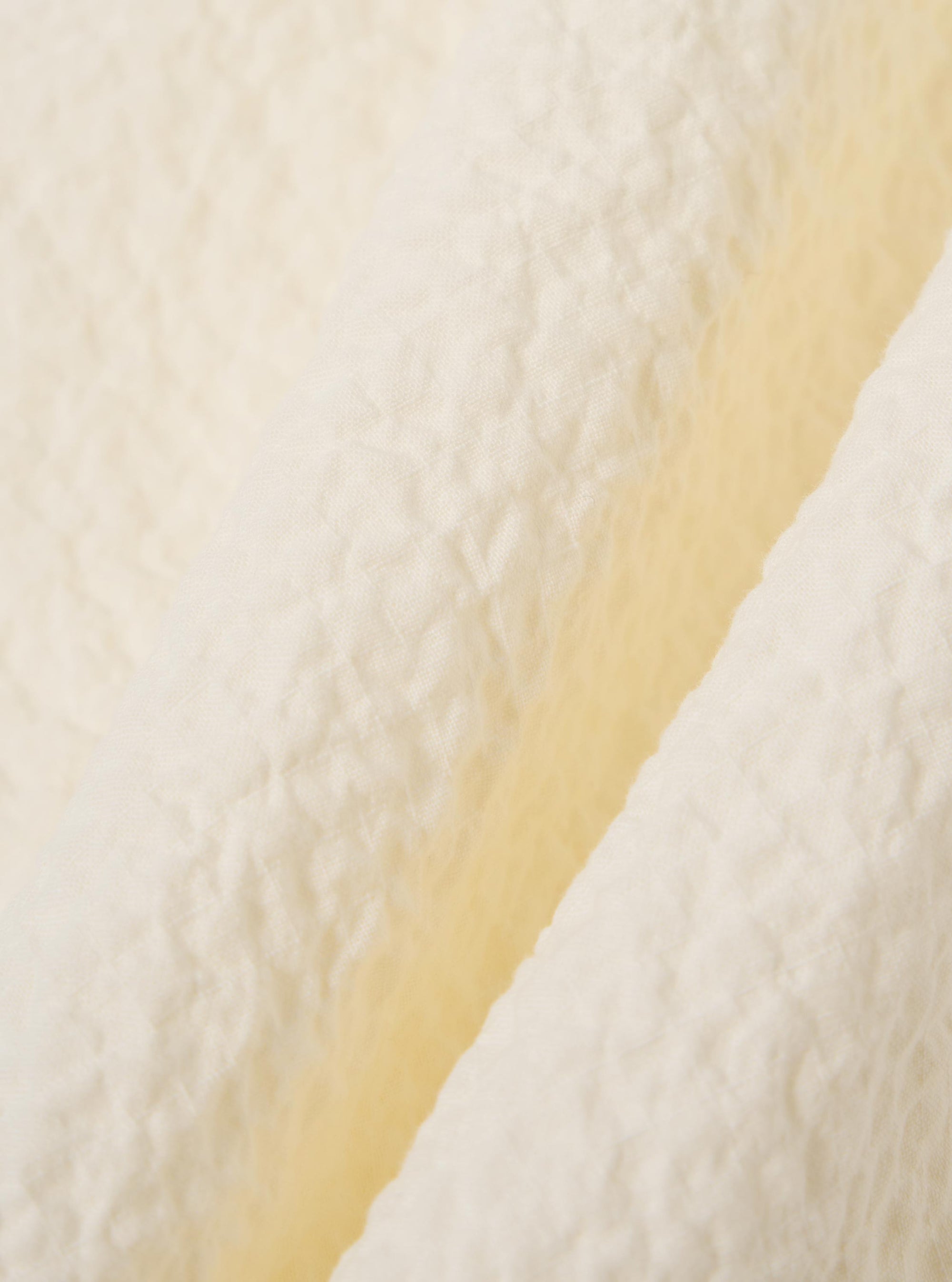 A close-up fabric swatch of the Ecru Arten Seersucker Road Shirt highlights the off-white ecru color and the distinct puckered texture of the organic cotton seersucker fabric. The material fills the frame, providing a detailed view of the textile's construction.