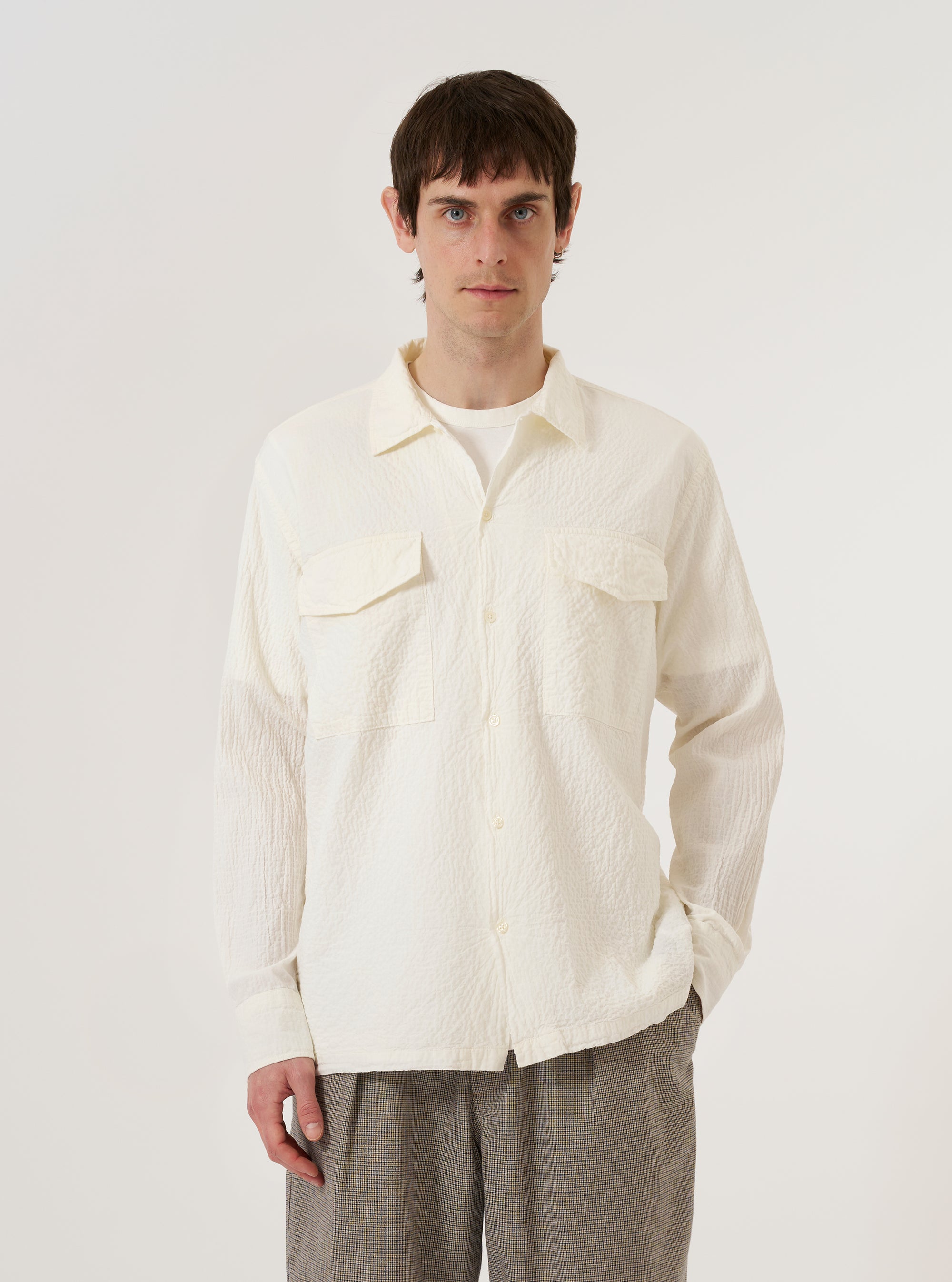 A model wears the Ecru Arten Seersucker Attendant Shirt over a white t-shirt, standing against a plain, neutral background. The shirt features a classic collar, a button-down front, and two large patch pockets at the waist. The ecru-colored organic cotton fabric has a distinct puckered seersucker texture.