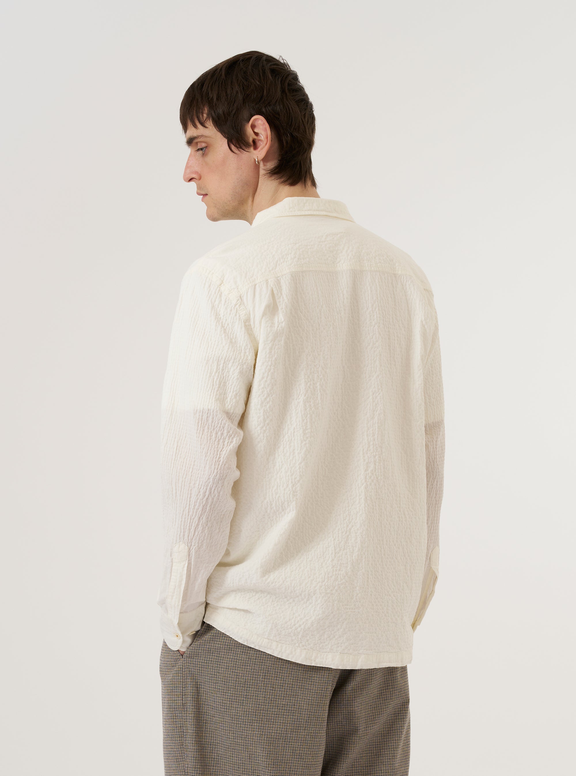 A model wears the Ecru Arten Seersucker Attendant Shirt, shown from a rear three-quarter angle against a plain, neutral studio background. The shirt features a utility-style construction with a straight hem and a simple collar. The ecru-colored organic cotton fabric highlights a prominent puckered seersucker texture.