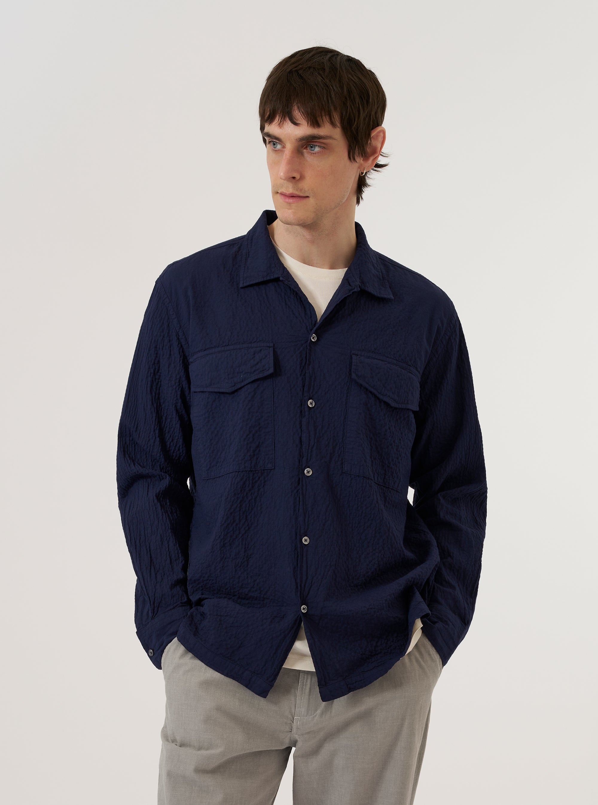 A model wears the Navy Arten Seersucker Attendant Shirt against a plain white background. The shirt features a loose fit, a one-piece collar, a central button closure, and two large oversized flap pockets on the chest. The deep navy fabric has a distinct puckered seersucker texture.