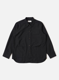 The Black Tropical Suiting Travail Overshirt is shown as a flat lay against a white background. The overshirt features a one-piece collar, a central button-down closure, and two large patch pockets at the waist. The lightweight black tropical suiting fabric has a smooth, minimal appearance.