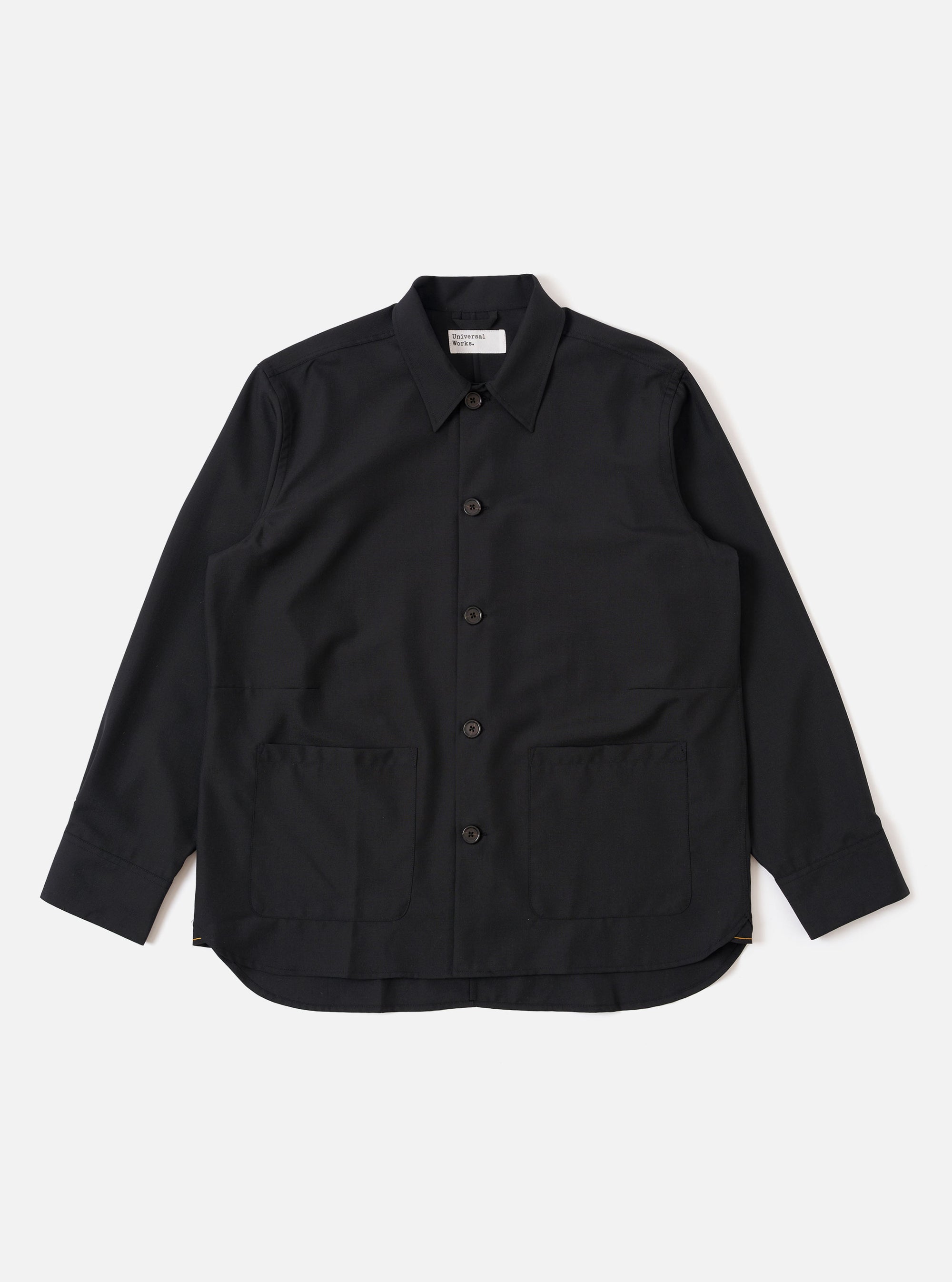 The Black Tropical Suiting Travail Overshirt is shown as a flat lay against a white background. The overshirt features a one-piece collar, a central button-down closure, and two large patch pockets at the waist. The lightweight black tropical suiting fabric has a smooth, minimal appearance.