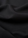 A close-up swatch image of the Black Tropical Suiting Travail Overshirt fabric. The image focuses on the textile detail, showing a solid black color with a smooth, lightweight weave characteristic of tropical suiting. The material fills the entire frame, highlighting its clean and professional finish.