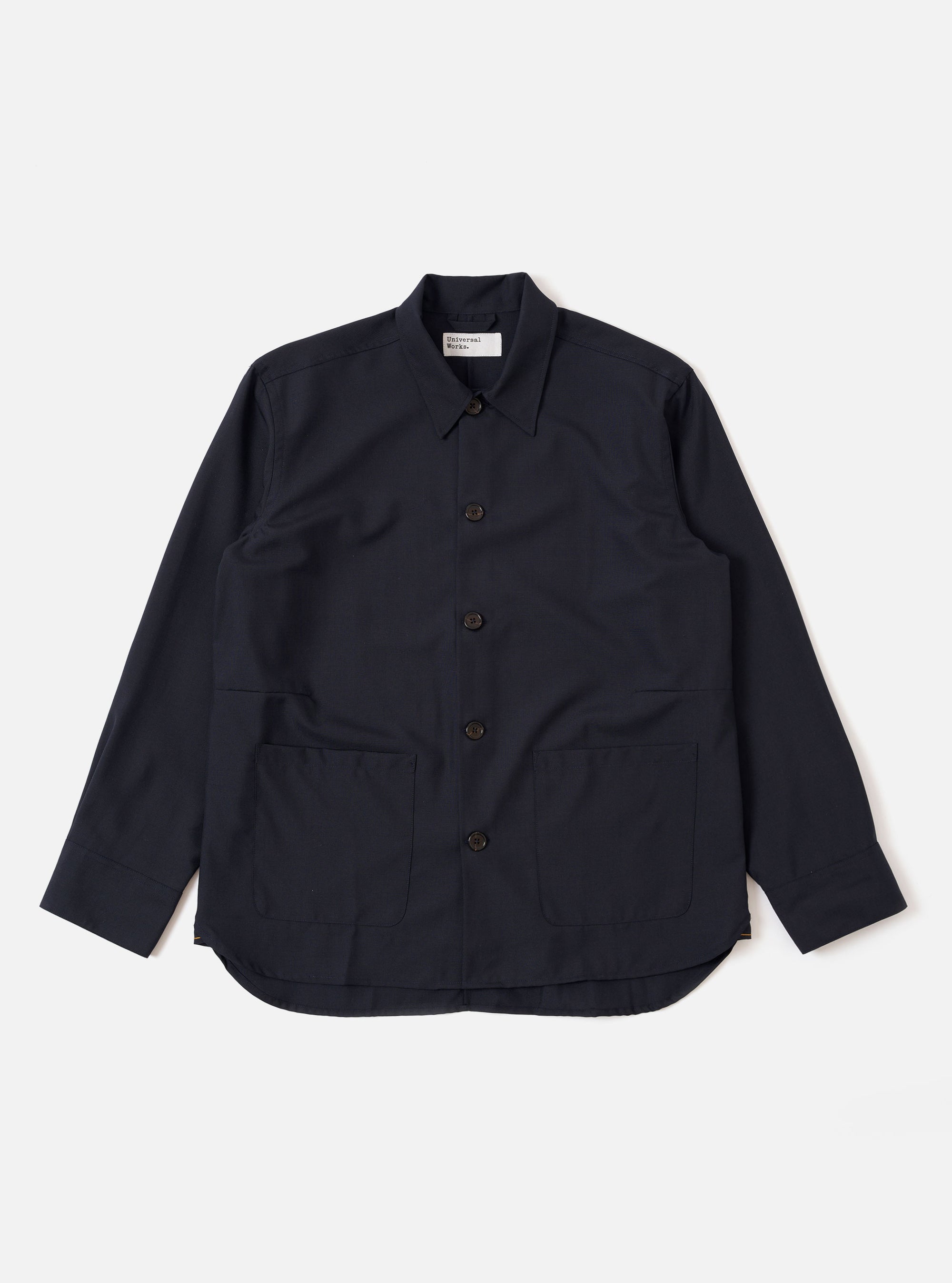 The Navy Tropical Suiting Travail Overshirt is shown as a flat lay against a white background. The garment features a single-piece collar, a button-through front, and two large patch pockets at the waist. It has adjustable button cuffs and a curved hem. The navy fabric is a lightweight tropical wool blend with a smooth, minimal appearance.