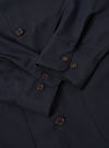 The Navy Tropical Suiting Travail Overshirt is shown in a detail view as a flat lay against a plain white background. The image focuses on the lower portion of the garment, highlighting the curved hem with a contrast fabric V-shaped insert at the side seam and the smooth, lightweight texture of the navy wool-blend fabric.