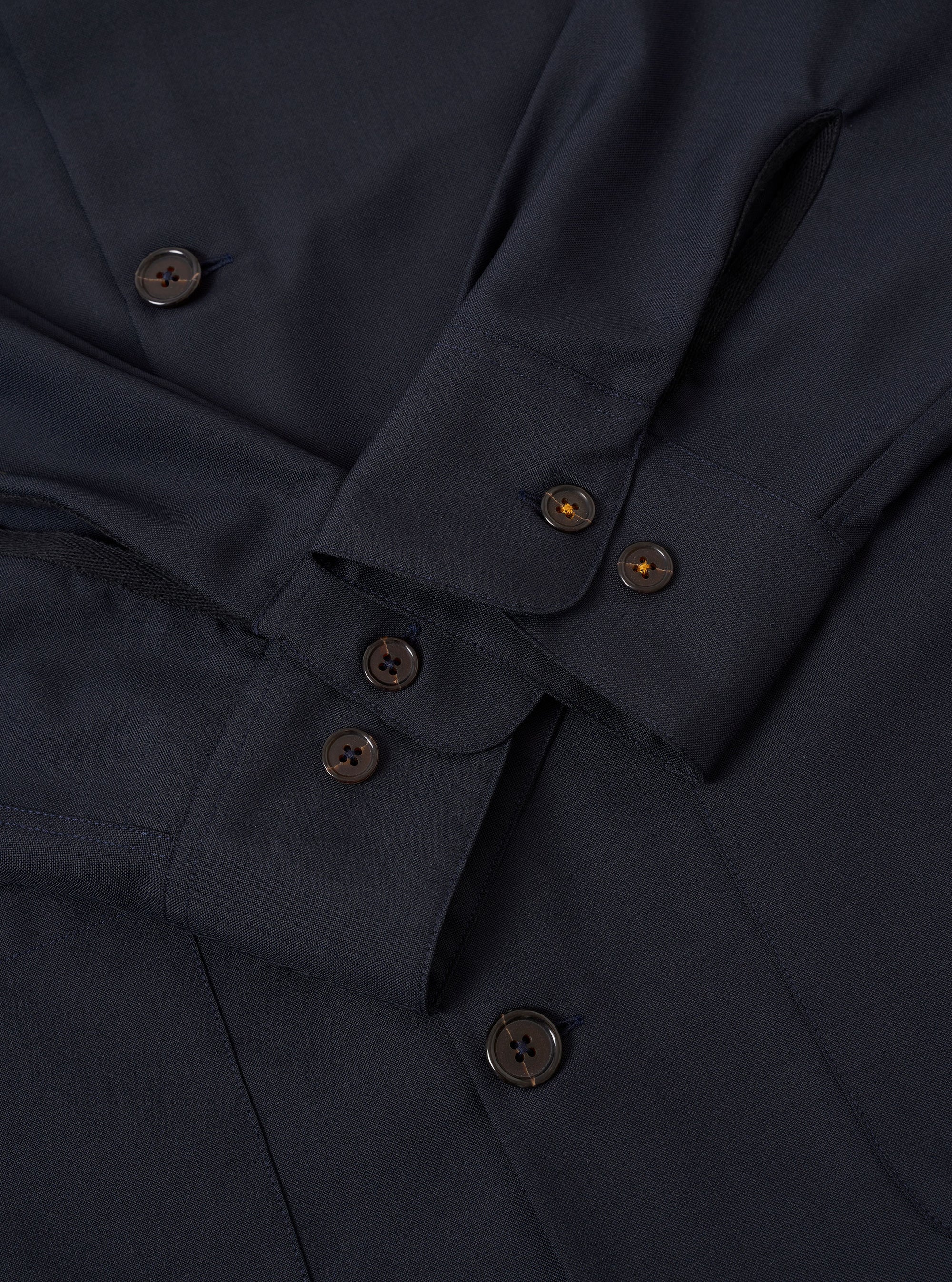 The Navy Tropical Suiting Travail Overshirt is shown in a detail view as a flat lay against a plain white background. The image focuses on the lower portion of the garment, highlighting the curved hem with a contrast fabric V-shaped insert at the side seam and the smooth, lightweight texture of the navy wool-blend fabric.