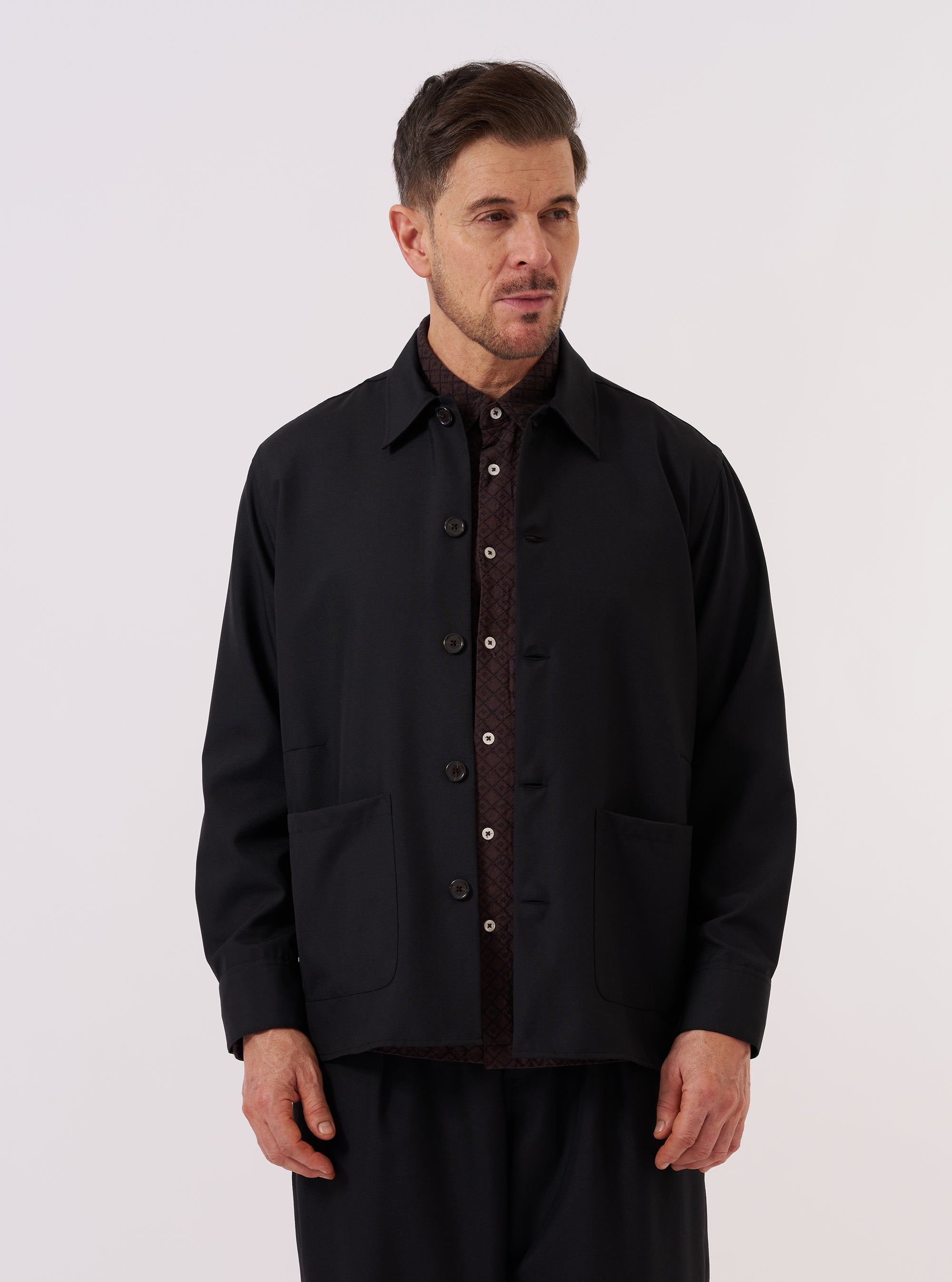 A model wears the Black Tropical Suiting Travail Overshirt over a light-colored shirt, standing against a plain, neutral background. The overshirt features a classic collar, a central button-down closure, and three prominent patch pockets on the front. The garment is made from a smooth black tropical suiting fabric.