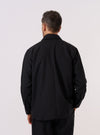 A model wears the Black Tropical Suiting Travail Overshirt, shown from the back against a plain white background. The overshirt features a structured collar, a straight hem, and a relaxed fit. The garment is constructed from a smooth black tropical suiting fabric with a lightweight, matte finish.