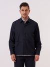 A model wears the Navy Tropical Suiting Travail Overshirt over a light-colored top, standing against a plain white background. The overshirt features a loose, unstructured fit, a one-piece collar, a central button-down front, and two large patch pockets at the waist. The navy blue tropical suiting fabric appears lightweight and smooth.