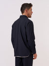 A model is wearing the Navy Tropical Suiting Travail Overshirt, shown from a side-angle view against a plain white background. The overshirt features a classic collar, a button-down front, and a large patch pocket visible on the side. The garment is constructed from a smooth, navy blue tropical suiting fabric.